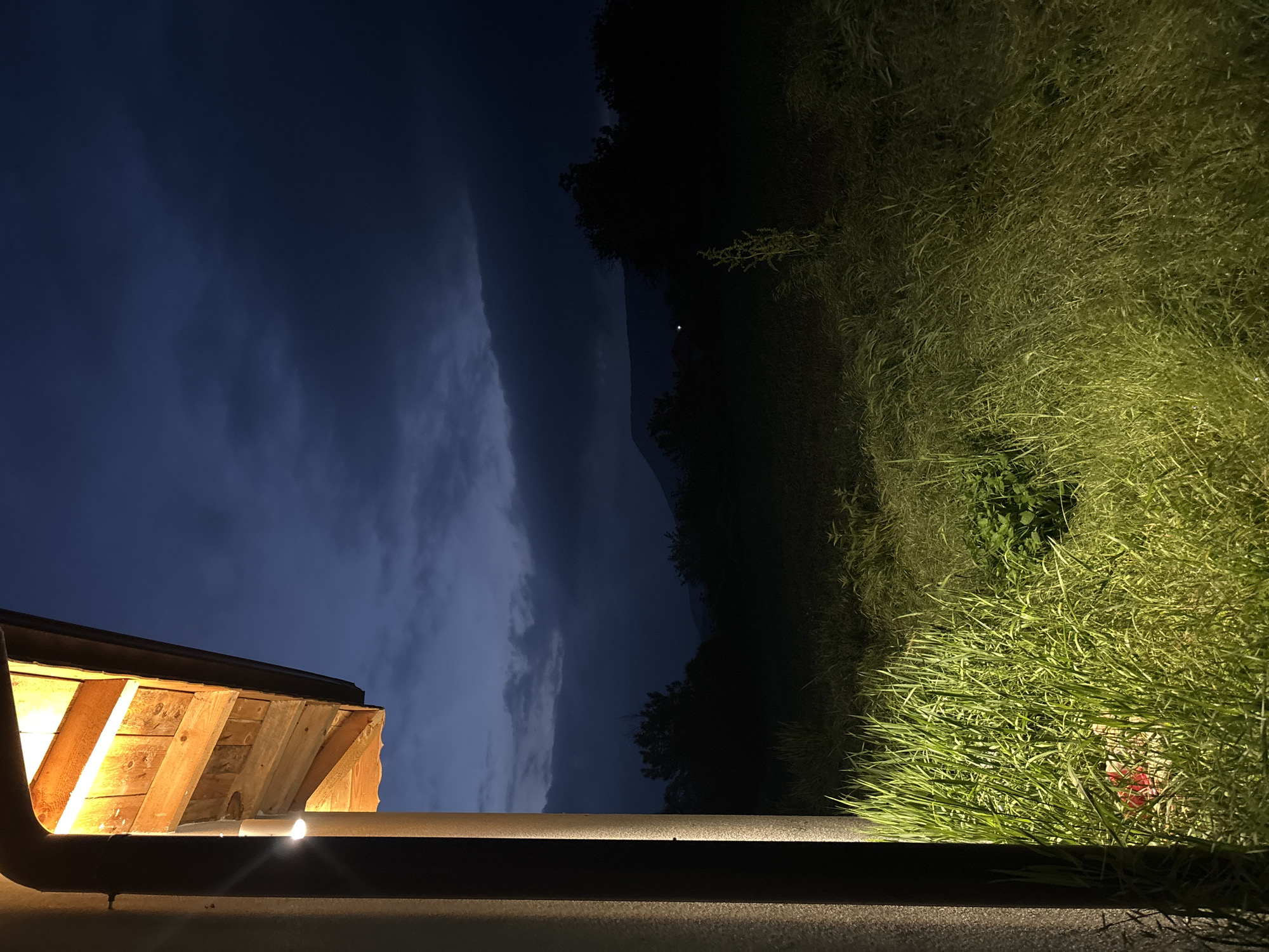 At night, a view from a balcony or porch shows a section of a wooden overhang illuminated by a warm light. A dark railing runs horizontally across the frame. Beyond the railing, there's tall, green grass, which appears to be lit by an unseen light source. In the distance, there's a dark line of trees or hills, and above that, a dark sky with a long, vertical cloud formation. The location is in the city of Unknown, Unknown.