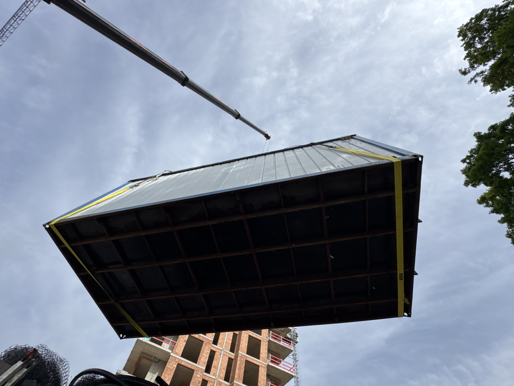 A large container with a dark bottom is suspended by yellow straps from a crane against a cloudy sky. The crane's arm extends from the top left. A building under construction with exposed brick and red railings is visible in the lower center. Trees are visible on the right side of the frame. A coiled pile of metal wiring is at the lower left corner. The location is identified as Unknown, Unknown.