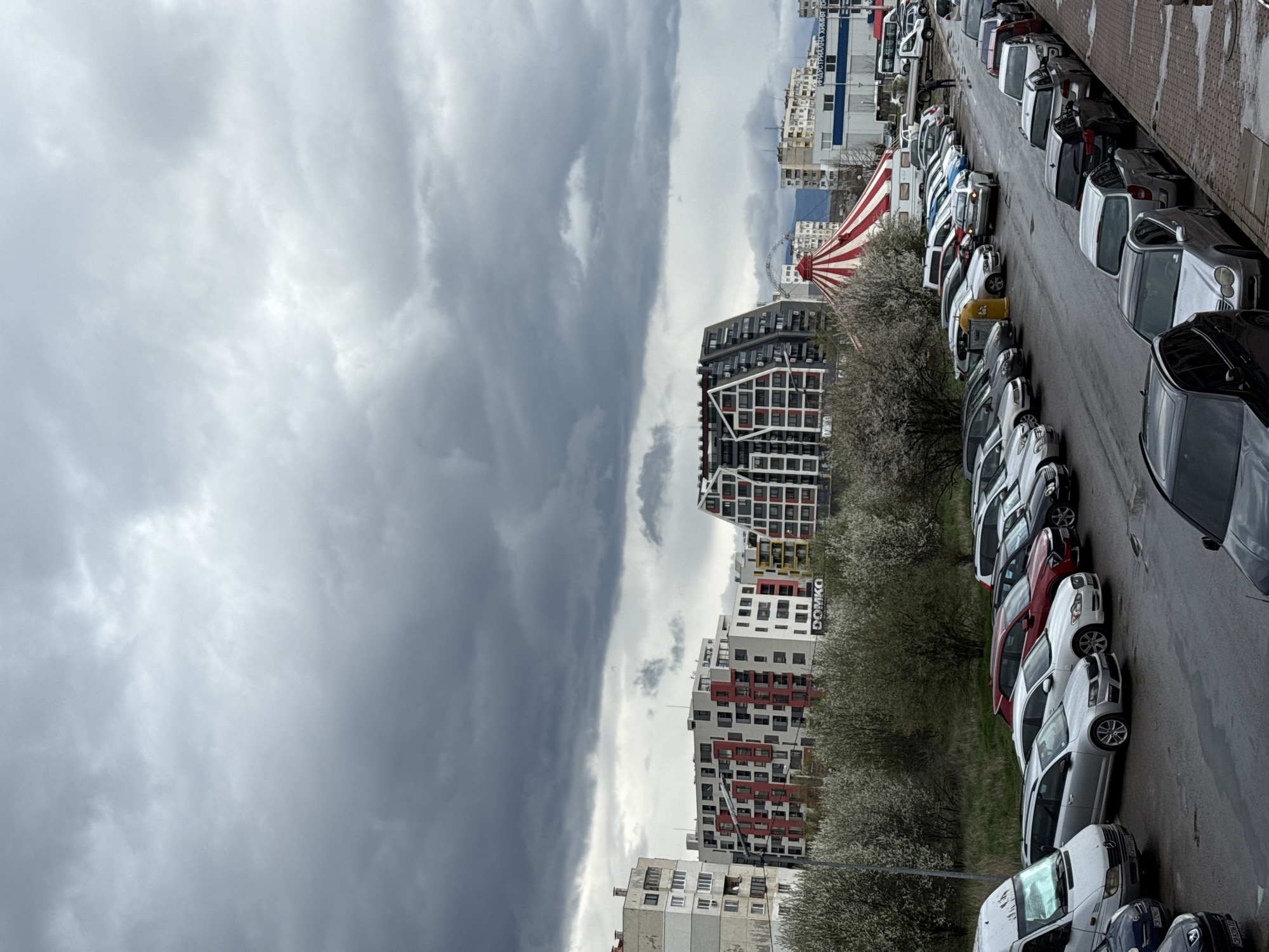 The image shows a street in Sofia, Bulgaria, lined with parked cars. The sky is overcast with dark, heavy clouds. On the left, there are several residential buildings of varying heights and designs. One building is distinguished by its red and black accents. Between the buildings and the cars are trees with white blossoms. A red and white striped tent-like structure is visible in the distance. The street is wet, indicating recent or ongoing rain.