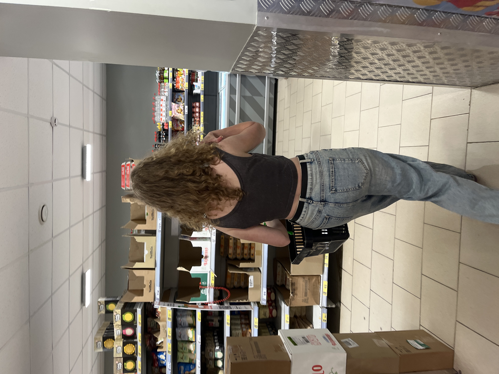 A person is standing in a grocery store aisle, viewed from behind. The person has curly brown hair and is wearing a dark grey tank top and light blue jeans. They are holding a black shopping basket in their right hand. The aisle is lined with shelves stocked with various products in boxes and bags. The floor is tiled with light-colored square tiles. In the background, other shelves with products are visible, along with a white ceiling with fluorescent lighting. The overall environment suggests a retail setting.