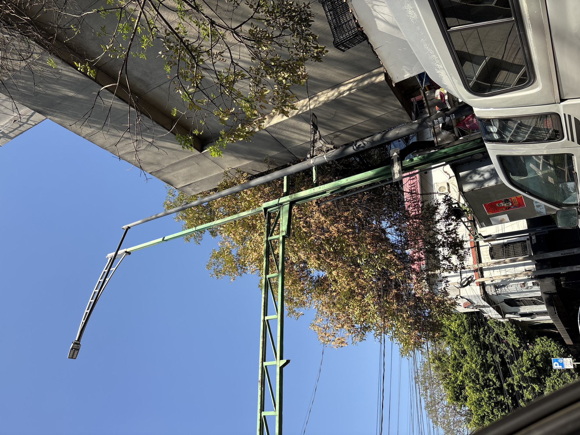 The image captures an urban street scene under a clear, bright blue sky, suggesting daytime with strong sunlight evident from the shadows cast. The primary feature is a large, sturdy green metal structure, possibly a gantry or support framework, with a lattice-like design. Extending from this structure is a long arm holding a modern LED street light.
To the right of the green structure, a white vehicle resembling a truck or bus is prominently visible, showing its front and side with dark windows and a headlight assembly. Behind it and beneath the green structure, another vehicle, possibly a garbage truck, can be partially seen.
Above the green structure and vehicles, there is a large concrete overhead structure, possibly an overpass or an elevated part of a building, from which branches of trees with sparse foliage and bare twigs hang down. More trees and dense green foliage are visible towards the right and bottom right of the image, some with light green leaves, others appearing autumnal or dry. Numerous black utility lines crisscross the frame, especially on the right side, extending from poles or buildings.
In the background, various buildings are discernible with windows, grates, and architectural details, some appearing residential or commercial. A small, blue sign with a white "P" symbol, commonly indicating parking, is partially visible near the bottom right amidst the foliage. Another red poster is visible on a building in the background but its text is illegible. No people are visible, and no specific event is taking place; the scene depicts a static moment in an urban environment.