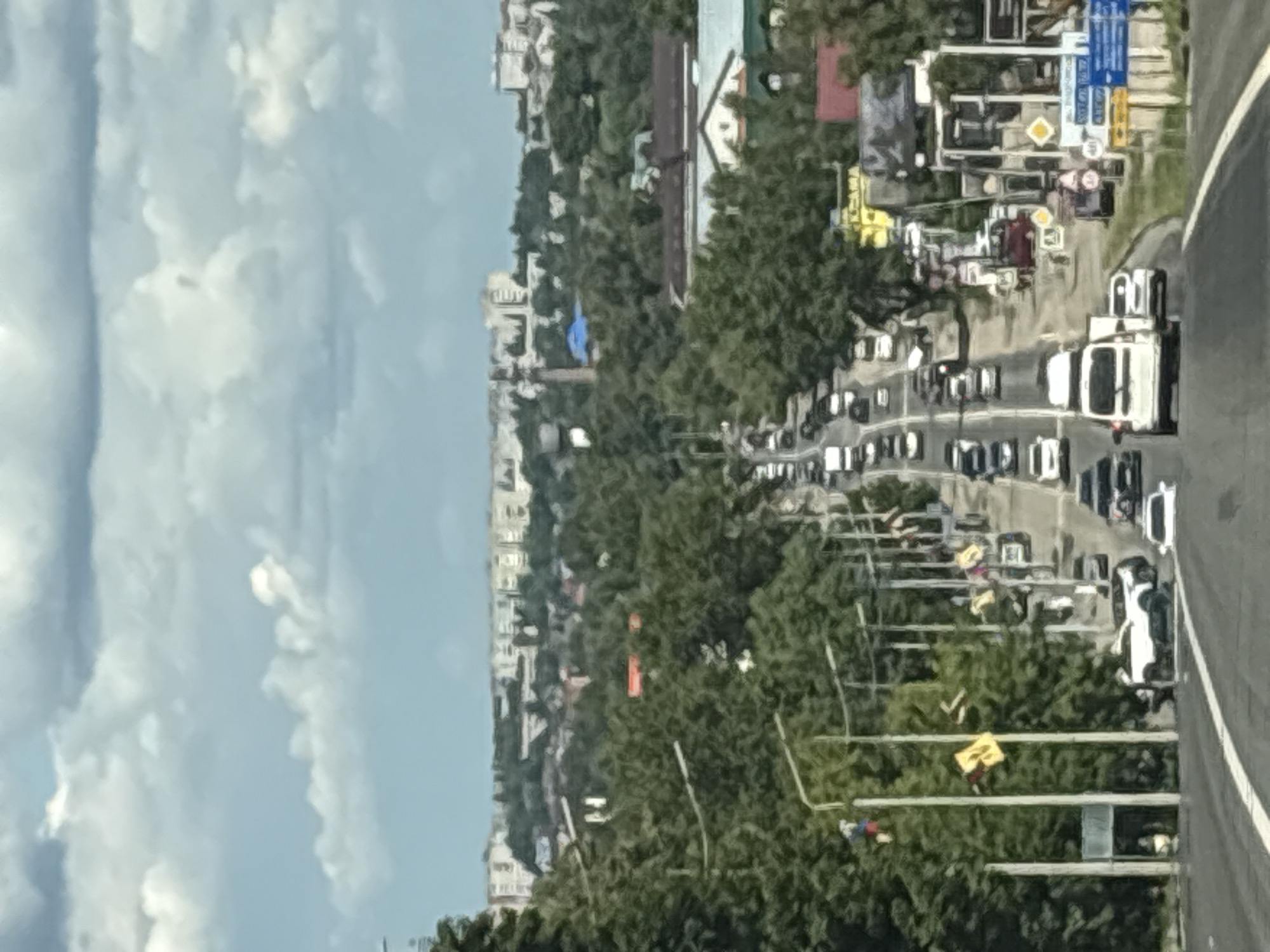 The image depicts a street scene in Pereslavl-Zalessky, Russia, viewed from a high vantage point, likely from a moving vehicle. The perspective is such that the road and buildings appear to be tilted. The scene is populated by numerous cars, indicating traffic. In the foreground, a white truck is prominent. Further back, rows of cars are visible on the road, suggesting a busy day. The surrounding environment is lush with trees, interspersed with apartment buildings of varying heights. The sky above is filled with white, fluffy clouds, suggesting fair weather. The presence of traffic lights and road signs indicates an urban or semi-urban setting. The overall impression is that of a typical day with moderate to heavy traffic in a Russian city. No discernible text is legible in the image.