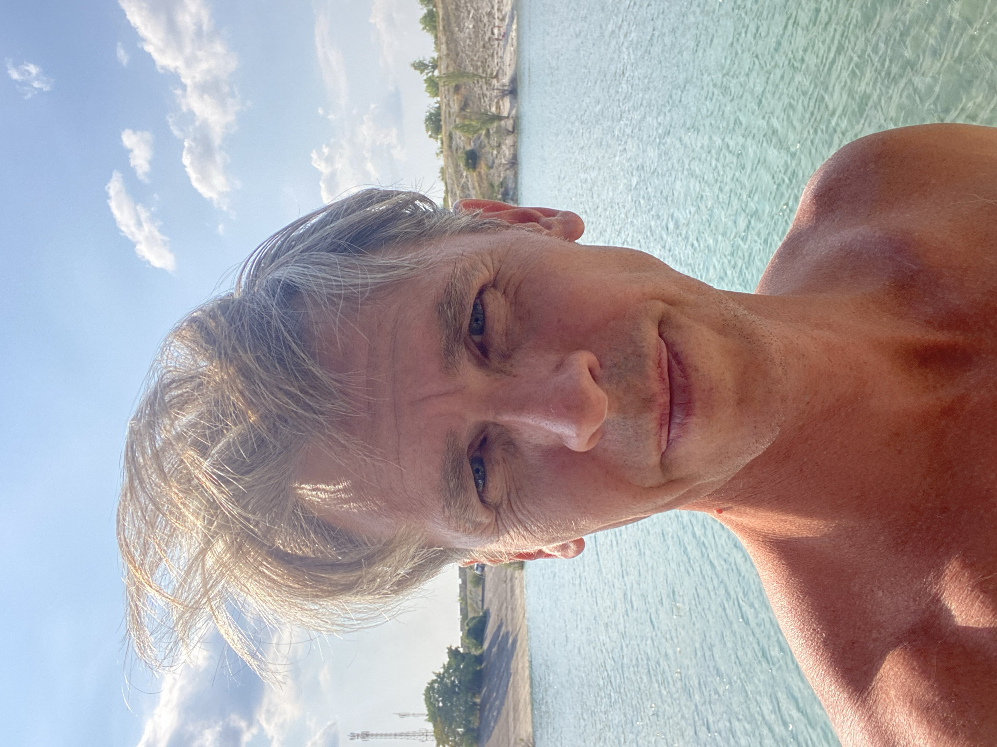 A middle-aged man with blondish hair is pictured from the chest up, looking directly at the camera. He is shirtless and his skin appears tanned. His hair is windswept and tousled, with some strands falling across his forehead. He is standing in front of a body of turquoise water, possibly a lake or quarry. In the background, there is a sandy embankment with sparse vegetation and a few trees. Two cell towers are visible on the left side of the embankment. The sky is blue with scattered white clouds. The man's expression is neutral. The location is specified as Kuybyshevskoye rural settlement, Ukraine, though this is a common name in multiple countries.