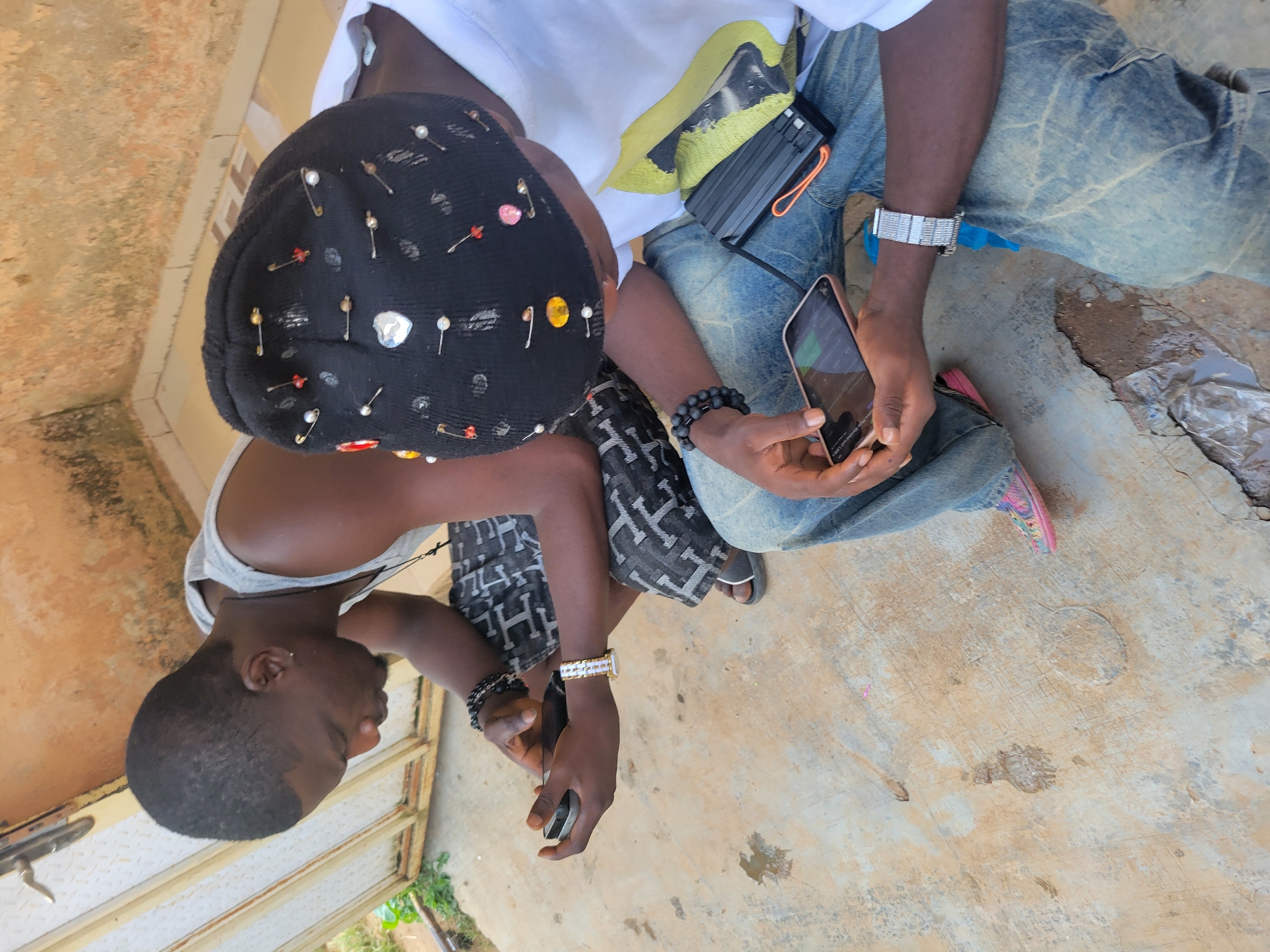 In an outdoor, urban setting, two individuals are seated on a concrete surface. The person on the left, a Black male with short hair, wears a gray tank top and patterned shorts, and is looking down at a smartphone in his left hand, which he holds horizontally. He has a black beaded bracelet on his left wrist and a silver-toned watch on his right wrist. He also appears to have a small earring in his left ear. The person on the right, whose face is obscured by a black beanie decorated with safety pins and beads, wears a white t-shirt and blue jeans. This individual is holding a smartphone in their right hand, also horizontally, and has a black beaded bracelet on their left wrist. A black object, possibly a portable charger, with an orange cord attached, rests on their lap. The background consists of a textured wall with a metal door frame. The concrete ground shows some discoloration and cracking.