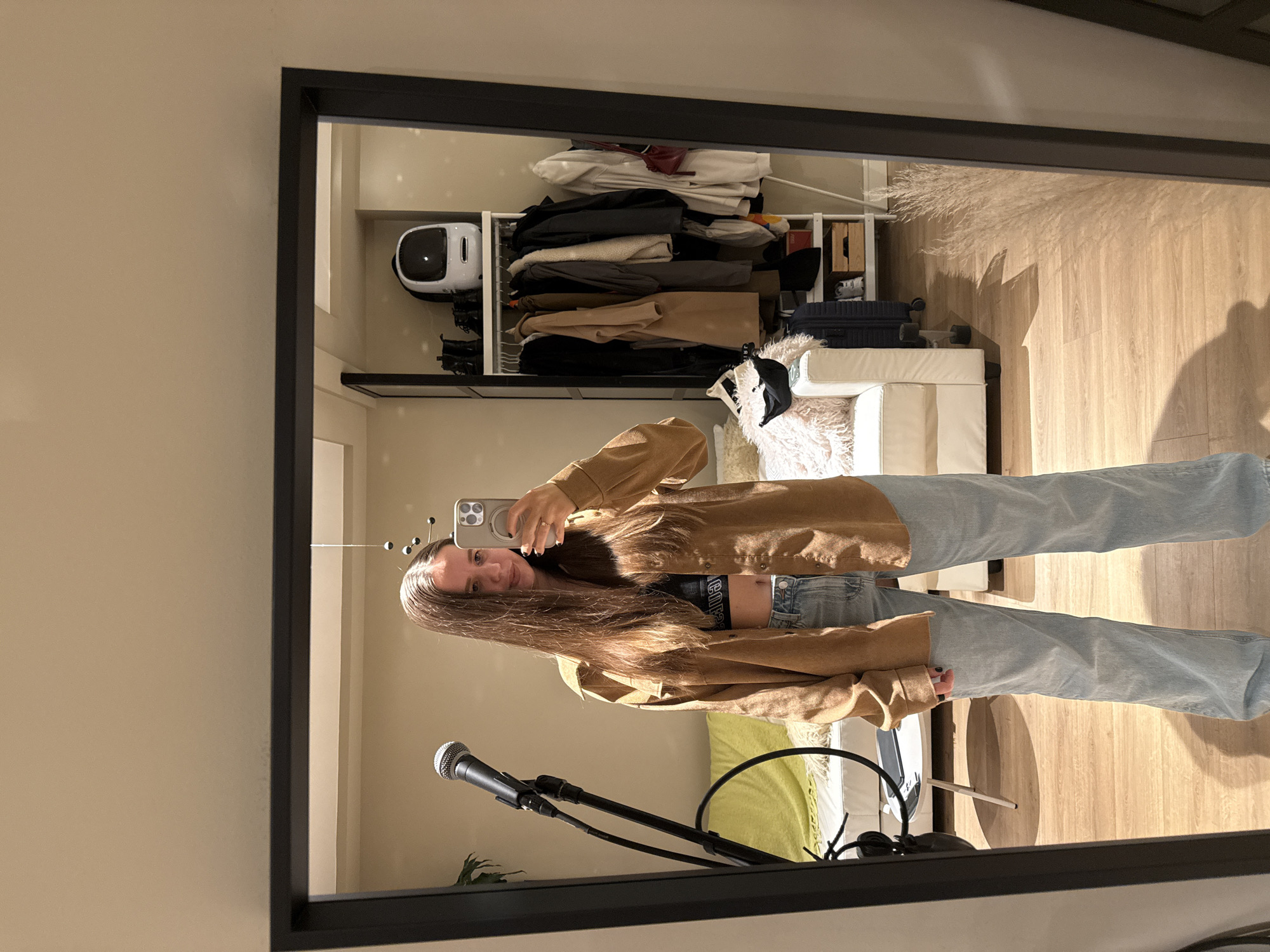 A young woman stands facing a mirror, taking a selfie with a smartphone. She is wearing a brown button-down shirt over a crop top, light blue jeans, and has long brown hair. The reflection shows a room with light-colored hardwood floors, a white wardrobe with clothes and accessories, including a white robot-shaped object on a shelf. There is also a white couch with fluffy cushions and decorative pampas grass. In front of the mirror, there is a microphone stand with a microphone attached. There is no clear indication this image was taken in Barcelona, Spain.