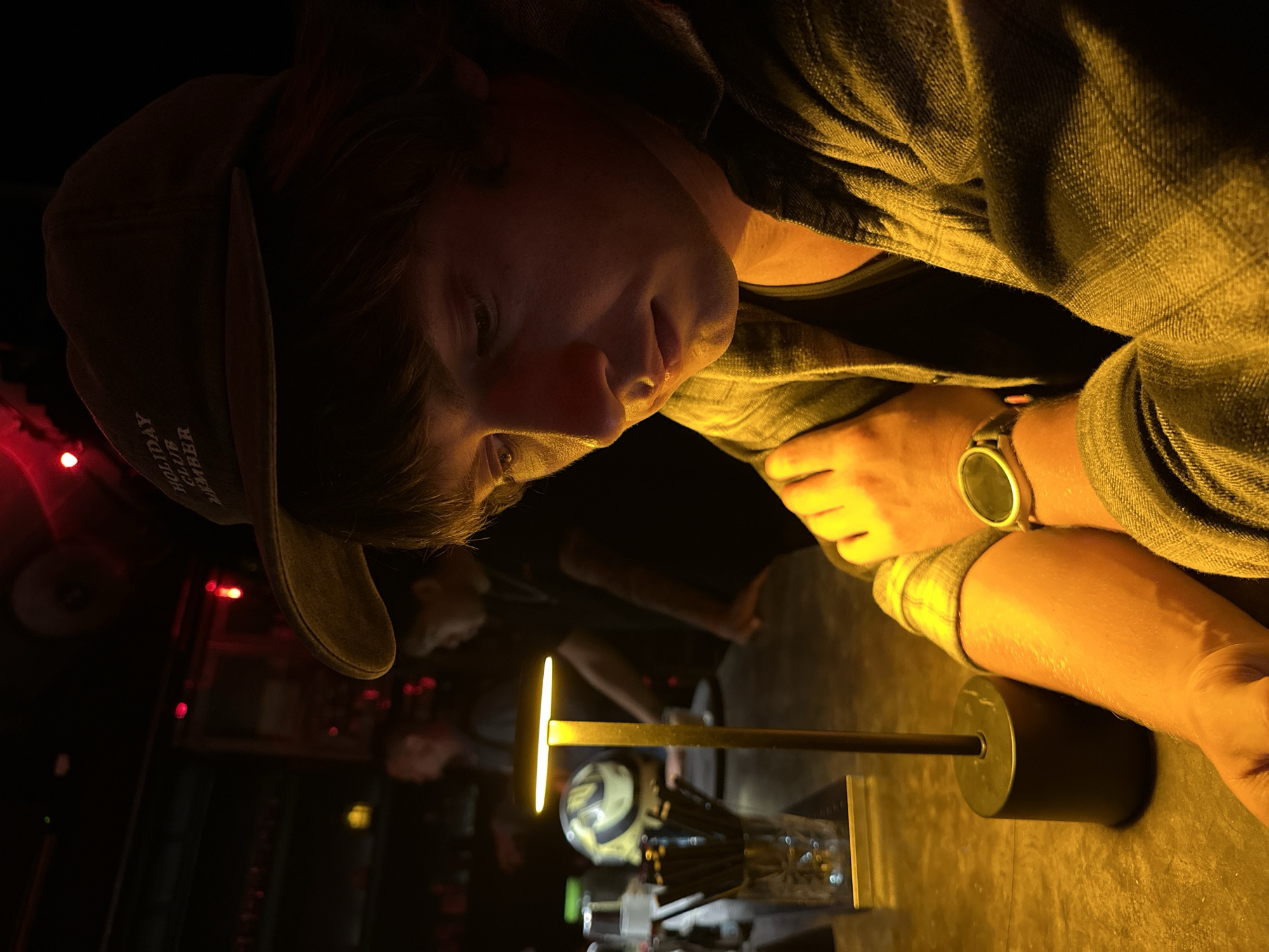 A man wearing a brown cap, plaid shirt, and a watch is seated at a table illuminated by a modern brass lamp. He is looking down towards the lamp, with his arm resting on the table. The background is dark, with faint outlines of other people and objects visible, along with red lights. The cap has the words "HOLIDAY CLUB AMBER" on it. The table is reflective, with a glass and various items visible near the lamp. The environment is dimly lit and seems to be a bar or restaurant. Based on the wording on the hat, it can be assumed that this image was taken in Canggu, Indonesia.
