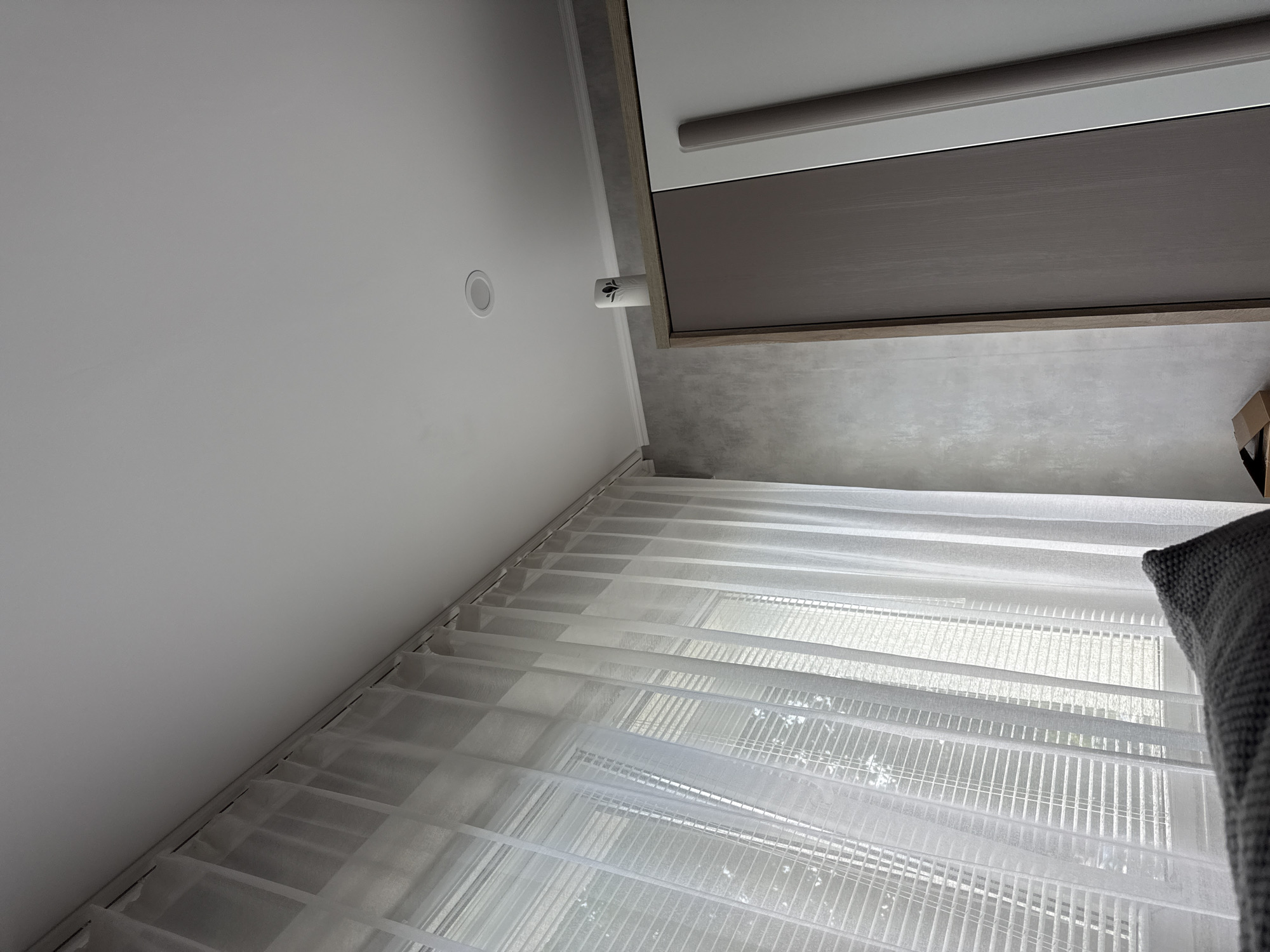 A low-angle interior shot depicts a room with white walls and a white ceiling containing a recessed light. A closed, gray roller shade is situated near the top right of the frame. Semi-transparent, white Venetian blinds cover a window, allowing diffuse light to filter through. A gray pillow rests near the edge of the frame in the lower right. The location is Lovech, Bulgaria.