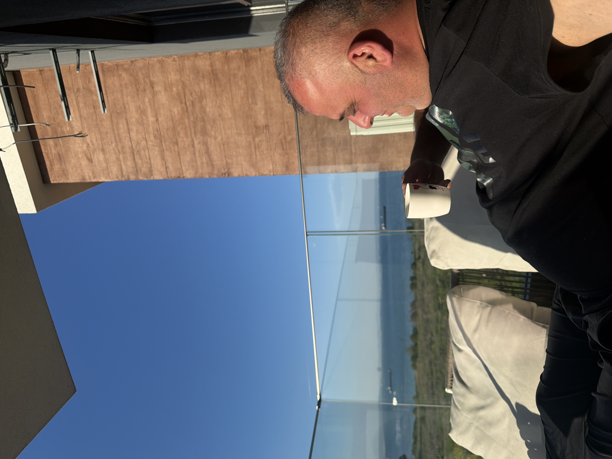 The image shows a man on a balcony holding a white mug. The man has a shaved head and is wearing a black t-shirt. The balcony has a glass railing, and the view beyond the railing includes water and land. The sky is blue. Based on the presence of the sea and buildings, this scene may be located in Burgas, Bulgaria.
