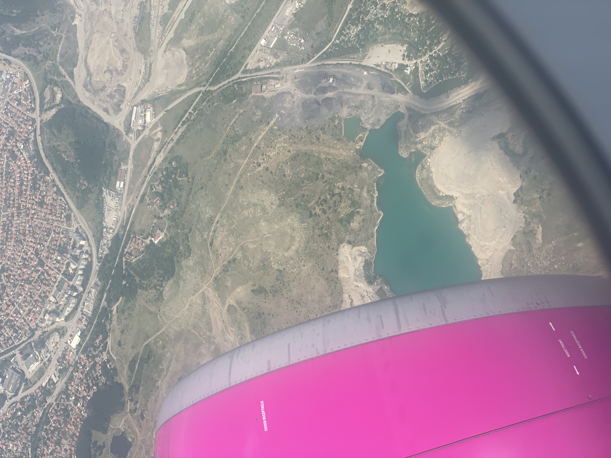An aerial view from an airplane window shows a landscape with a body of turquoise water, a mine or quarry, and a residential area with red-tiled roofs. A portion of the airplane's pink engine cowling is visible in the foreground, with the text "COVER RECEPTACLE" and "HOIST POINT" printed on it. The residential area appears to be a dense collection of houses. The mine or quarry area features mounds of earth and what appears to be coal or dark material. Roads wind through the landscape, connecting different sections. The overall scene depicts a somewhat industrial or developed area adjacent to a natural landscape.
