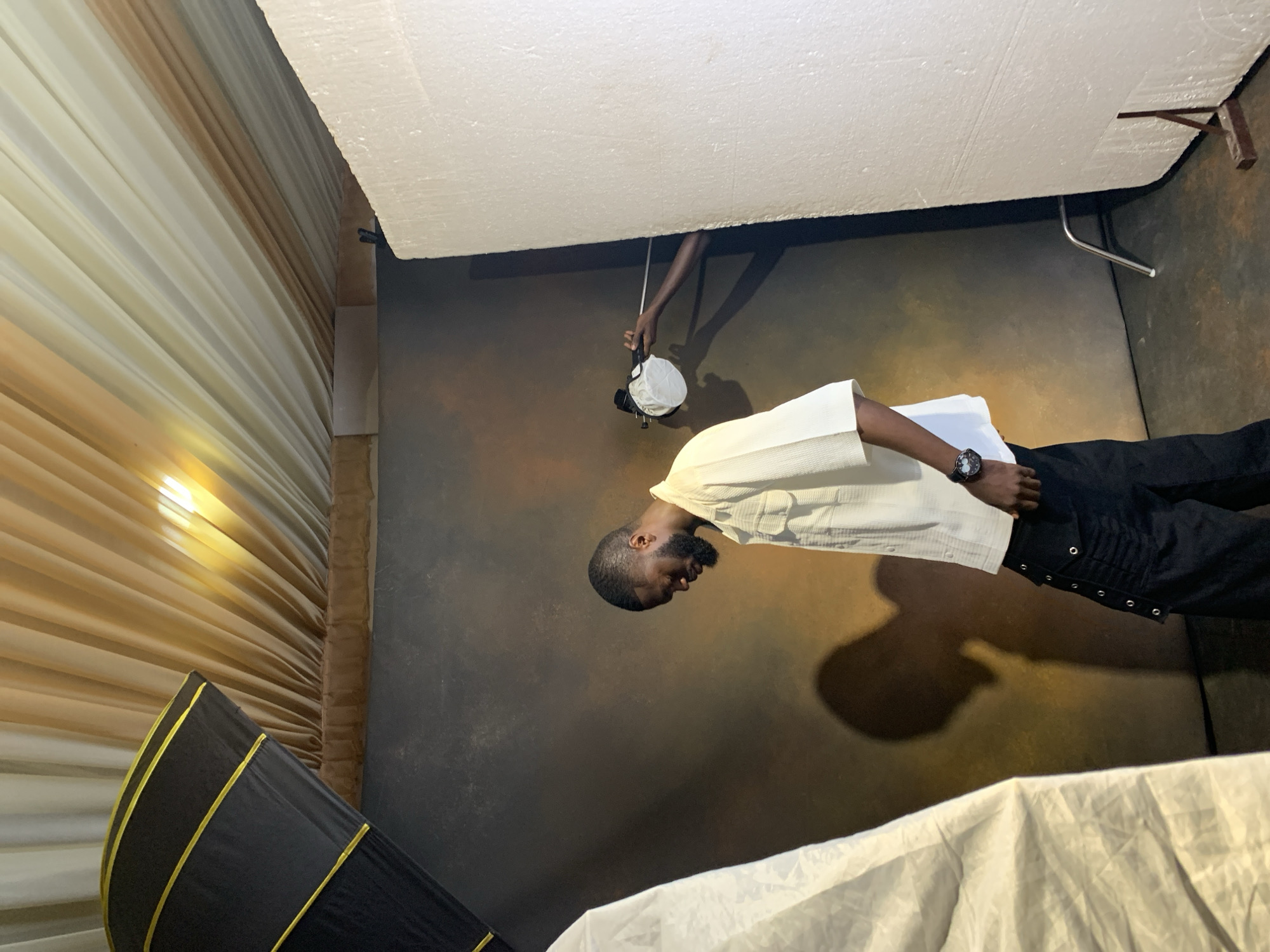 In a studio setting in Barakin Sabawa, Nigeria, a man is being photographed. He is wearing a cream-colored collared shirt and black pants, with a watch on his left wrist. The studio has a brown, textured backdrop. A large, white piece of styrofoam is positioned above the man, and a hand is visible holding a light modifier or camera accessory, pointing it towards the man. To the left of the frame, a black and yellow photography umbrella is partially visible. The lighting suggests an indoor shoot, likely during the daytime. The man's expression is neutral as he looks downwards. The scene is arranged for a professional photoshoot, indicated by the studio equipment.