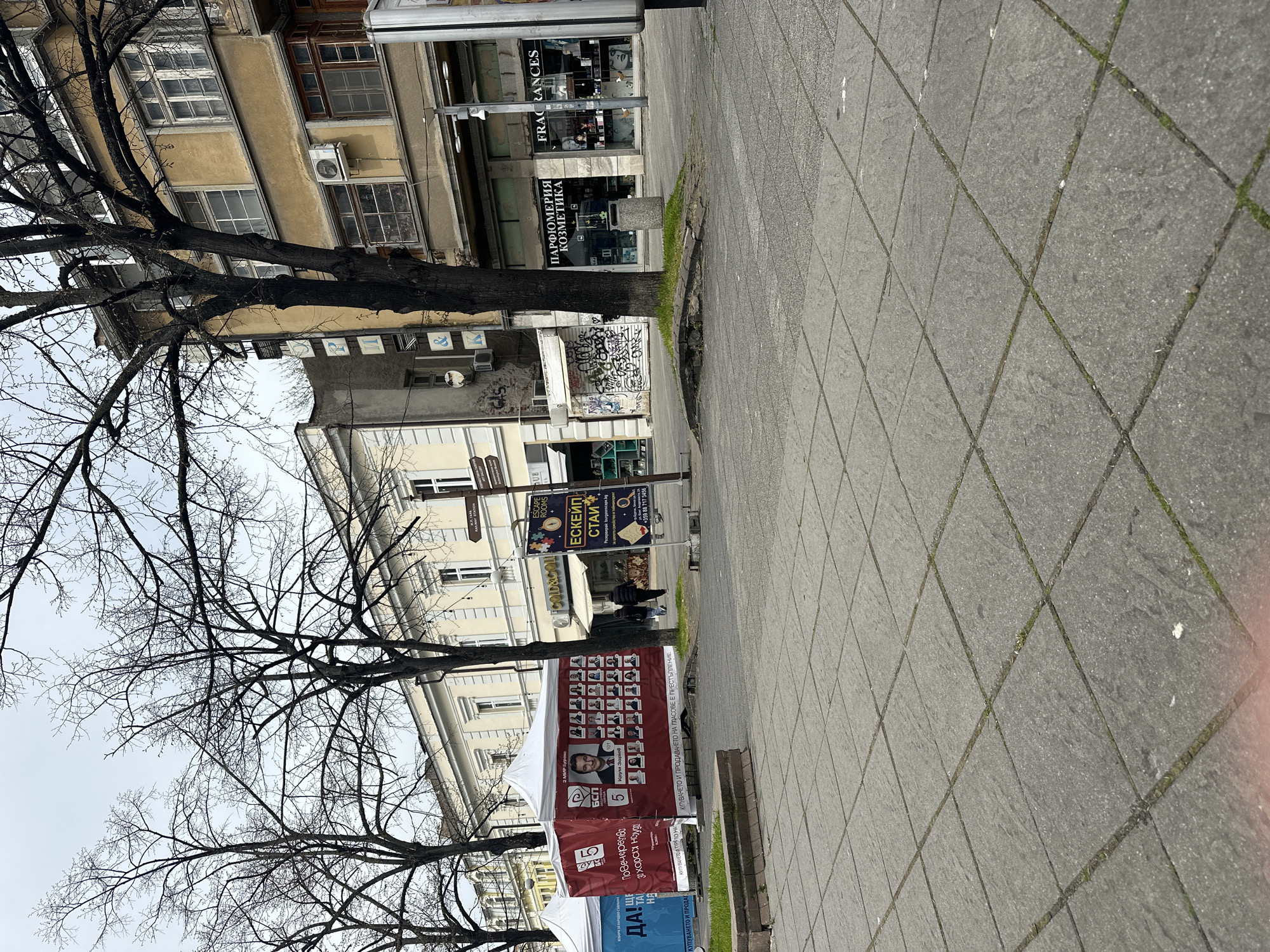 The image depicts a street scene in Burgas, Bulgaria. The camera angle is slightly off-kilter, showing a tiled sidewalk in the foreground, stretching into the distance. A bare tree, devoid of leaves, frames the left side of the image. Several buildings of varying heights and architectural styles line the street. Commercial establishments, including a fragrance store and an escape room business, are visible. Political posters are displayed on tents, suggesting an election period. Two people are walking in the distance. The sky is overcast.