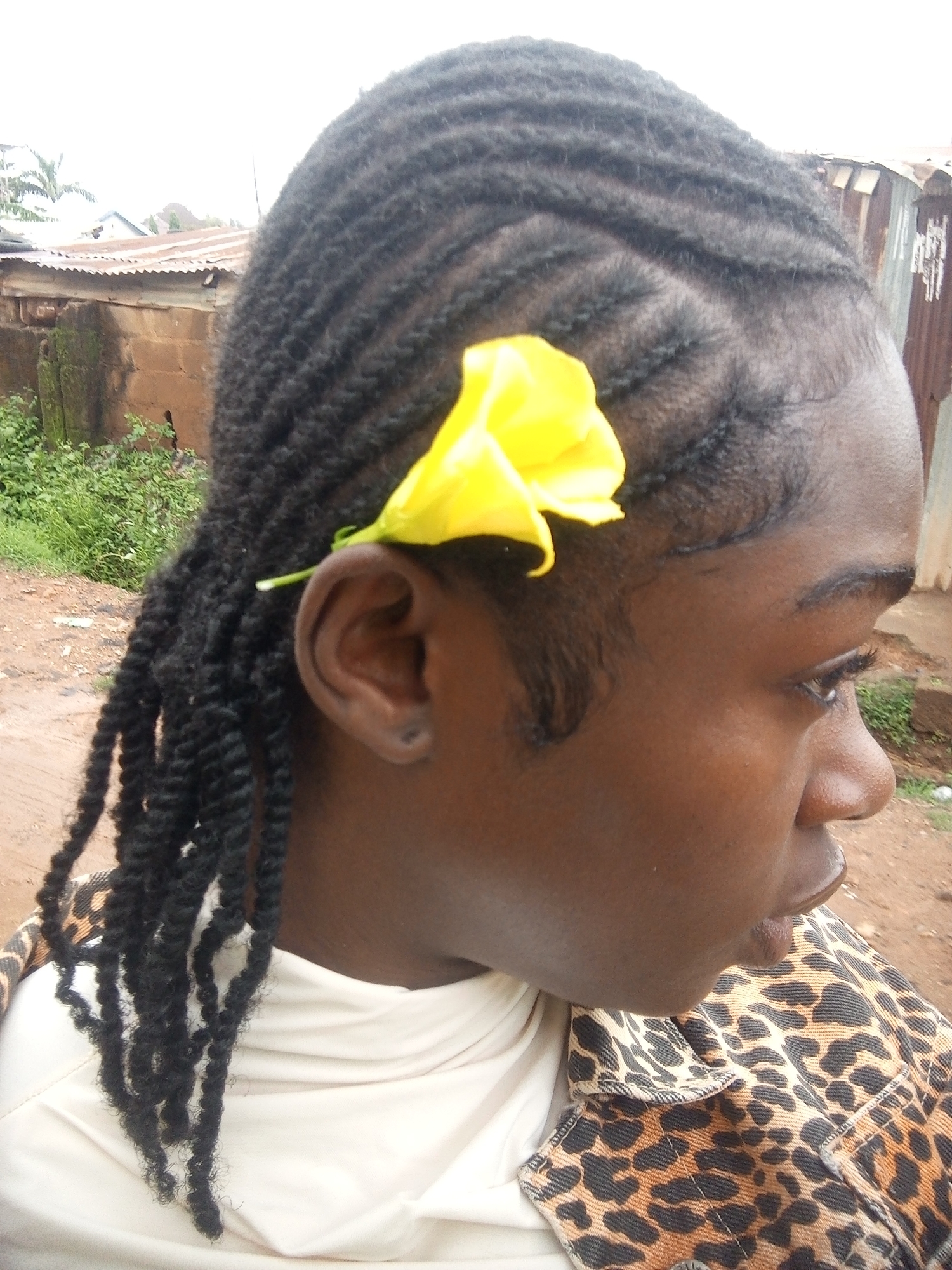 The image is a close-up, profile shot of a young Black female from the neck up. She is outdoors, with a blurry background suggesting a rural or semi-urban setting. Her hair is styled in cornrows that transition into thick, twisted braids. A bright yellow flower is tucked behind her ear, against her scalp. She is wearing a light-colored garment underneath a leopard-print jacket. The background shows a rustic corrugated metal structure and some greenery. The overall impression is of a casual, everyday moment captured in what appears to be a developing area. There is no information in the image to suggest the location is specifically Rafin Par, Nigeria, or any other city for that matter.