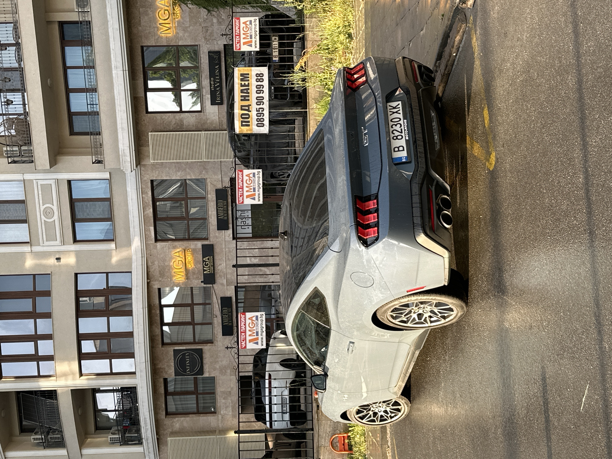 The image shows a gray Ford Mustang parked on the street next to a building. The building has a beige facade with multiple windows and several business signs, including "MGA Group", "Studio Irina Velina", "Ambero Beauty Lounge" and "Infinity Realty for NEE". There is also a sign that reads "ПОД НАЕМ 0895 90 99 88" (FOR RENT 0895 90 99 88). Another car, possibly a black SUV, is parked behind a fenced area. License plate on the Mustang reads "B 8230 XK" (Sofia, Bulgaria). The sign indicates that this is a private parking area. The setting appears to be an urban environment, and based on the Cyrillic text on the signs, the location is likely in Bulgaria, most likely the city of Varna.