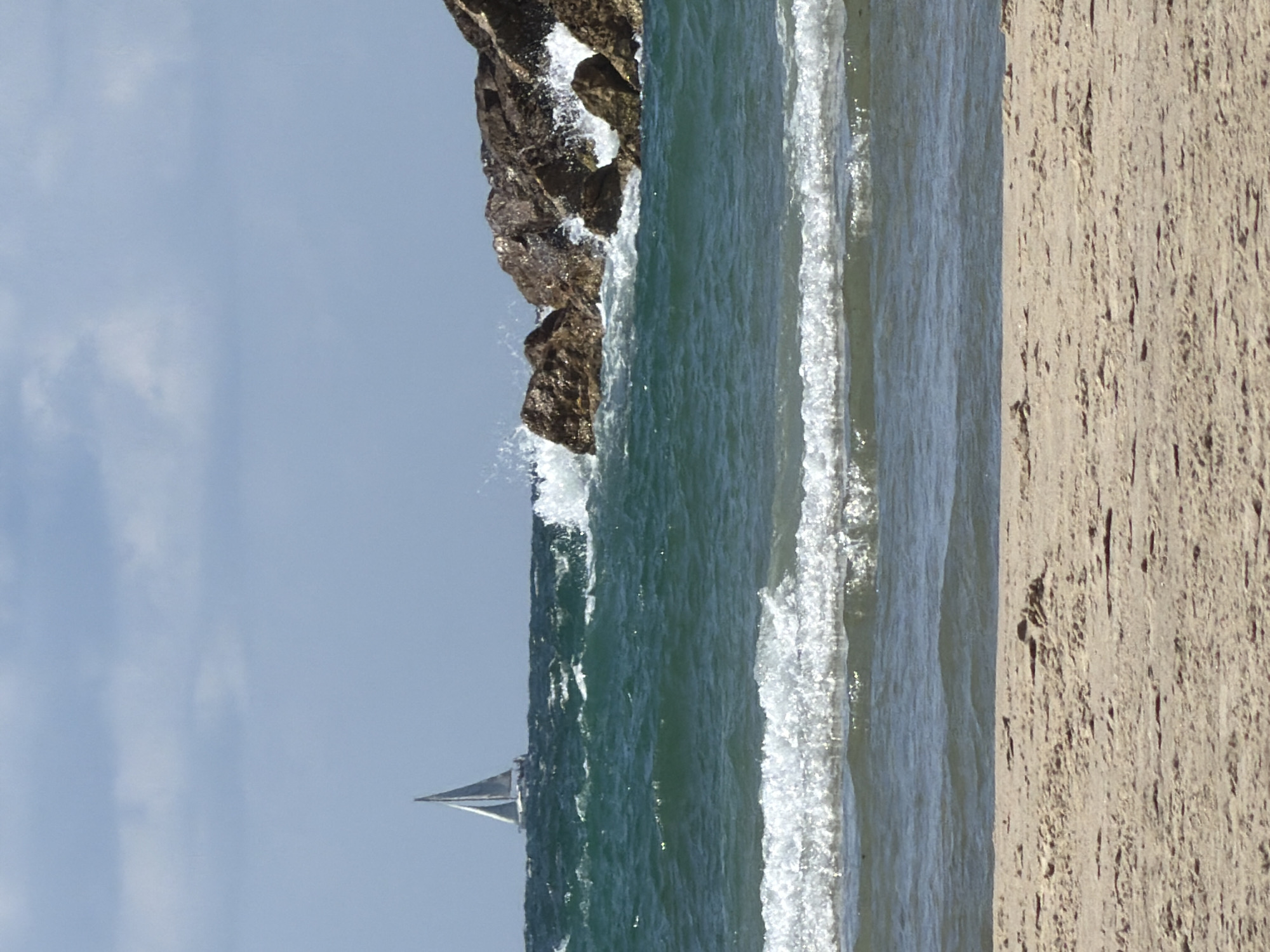 The image depicts a coastal scene, possibly near Tel-Aviv, Israel, but without verifiable certainty. A sandy beach occupies the right portion of the frame. The sea extends into the center, with waves breaking near a rocky outcrop. A sailboat is visible in the water, set against a pale blue sky. The image is presented in a vertical orientation.