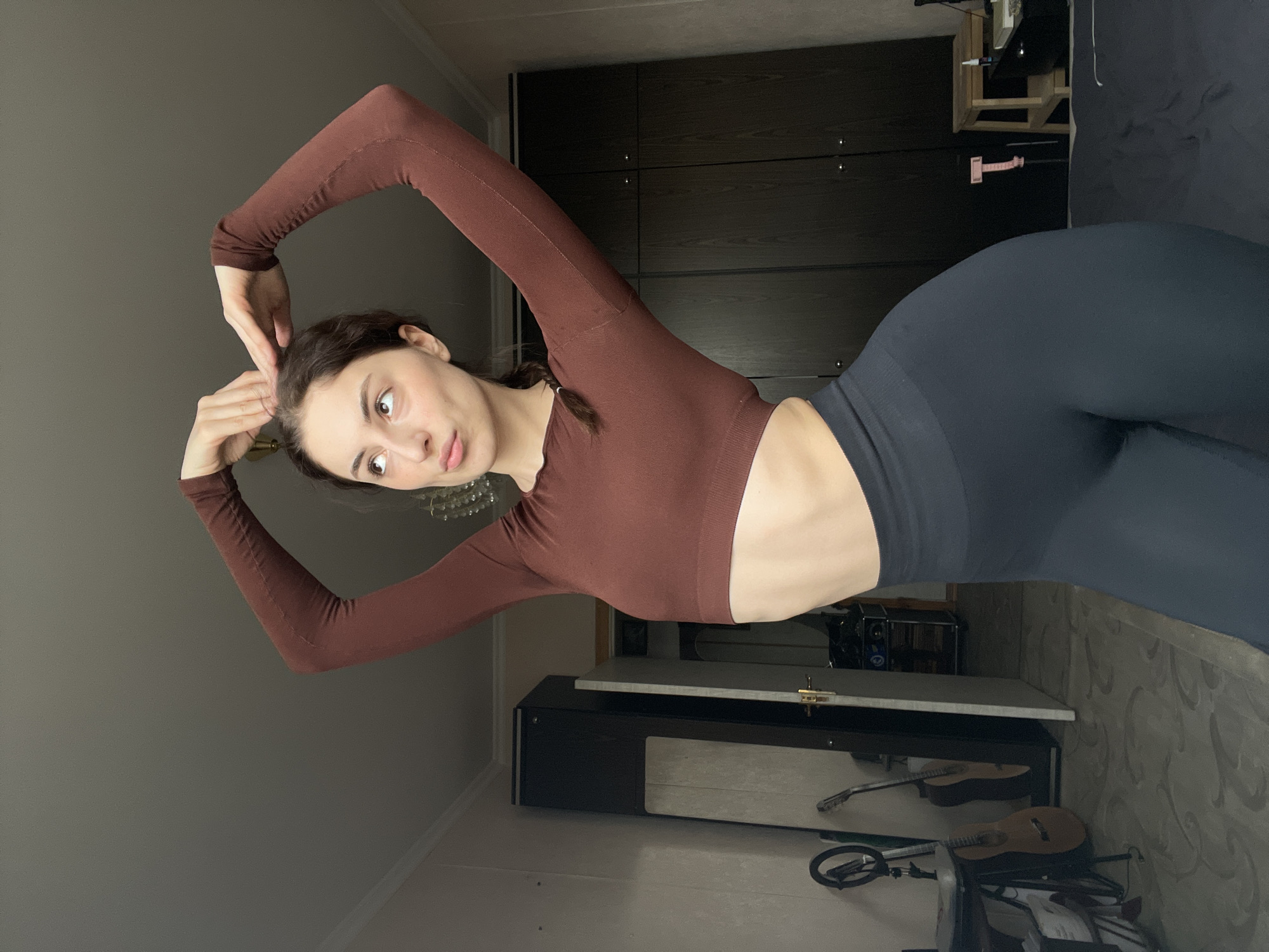A young woman is posing indoors. She is wearing a long-sleeved brown crop top and dark leggings. She has dark hair, pulled back in a partial braid. Her hands are raised near her head. The background includes dark furniture resembling a wardrobe, a shelf with items, and musical instruments including a guitar. The location cannot be determined from the image.