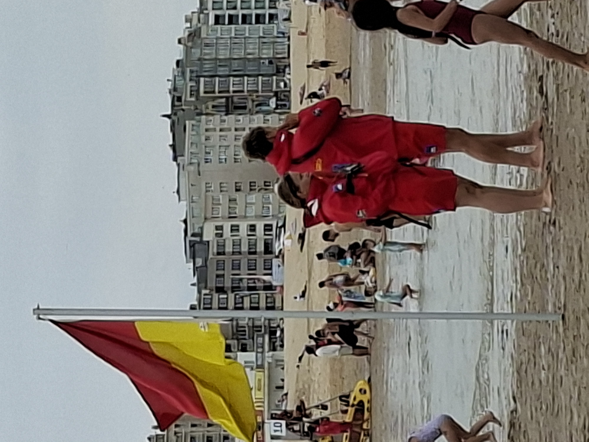 The image depicts a crowded beach scene in Ostend, Belgium, likely during daytime. In the foreground, two lifeguards, a man and a woman, are standing on the sand, both wearing red jackets over red shorts. The woman is looking to her left, while the man is looking to his right, with his hand near his face. To the right, a young girl in a maroon swimsuit walks past them. Behind the lifeguards, a red and yellow flag waves from a flagpole. The beach is populated with many people, some wading in the water, others sitting or walking on the sand. In the background, there is a line of multi-story buildings, characteristic of a seaside resort town. The sky is overcast, suggesting potentially cloudy weather. A yellow lifeguard vehicle is visible to the left of the frame. The scene captures a typical summer day at a busy beach.
