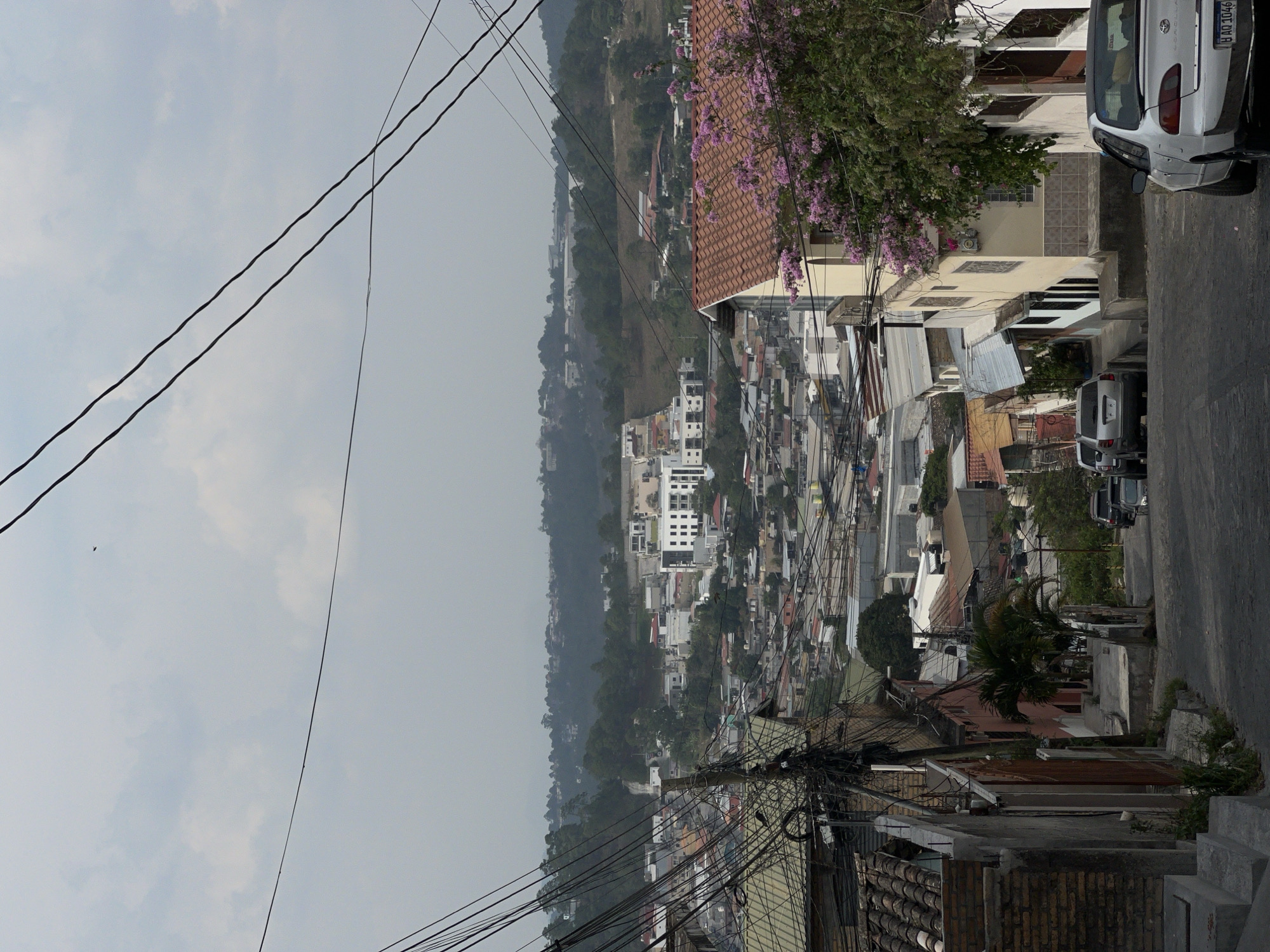 The image shows a cityscape in Santa Rosa de Copán, Honduras. It is taken from an elevated position looking down at buildings and houses of varying sizes and designs. A valley stretches out in the background under an overcast sky. Overhead power lines crisscross the foreground of the image. Along the right side of the frame is a street with parked cars, including a white Toyota with license plate HAD 1046. Vegetation, including trees with pink flowers, is visible. The general atmosphere is one of urban density and a somewhat hazy environment.