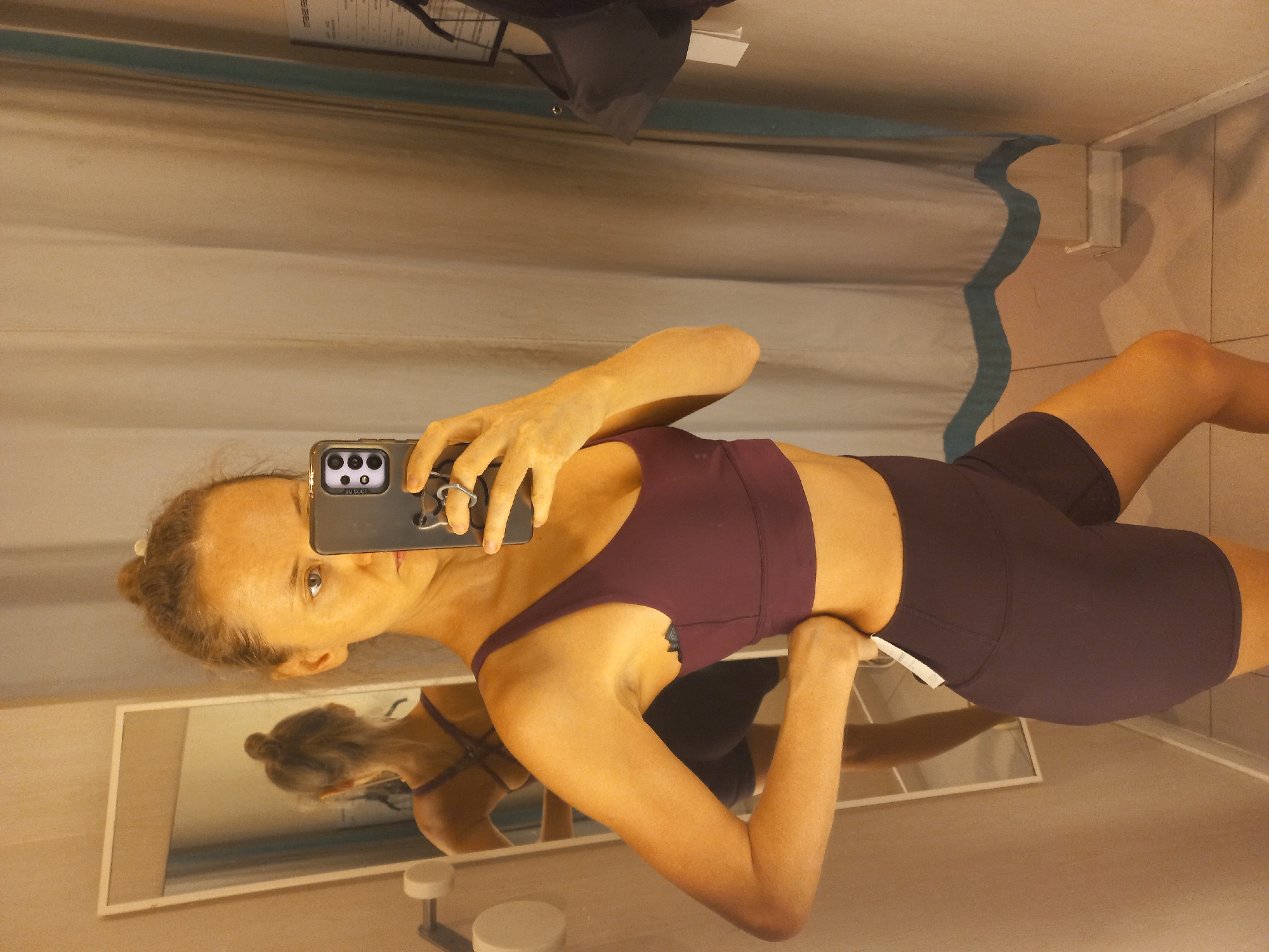 A woman is taking a selfie in what appears to be a clothing store dressing room. She is wearing a maroon-colored sports bra and matching shorts. She is holding a black smartphone in her right hand to take the photo. The room has beige walls, a mirror, and a white curtain with a blue design on the top edge. A black garment is partially visible hanging above the curtain.
Location cannot be determined solely from the image.