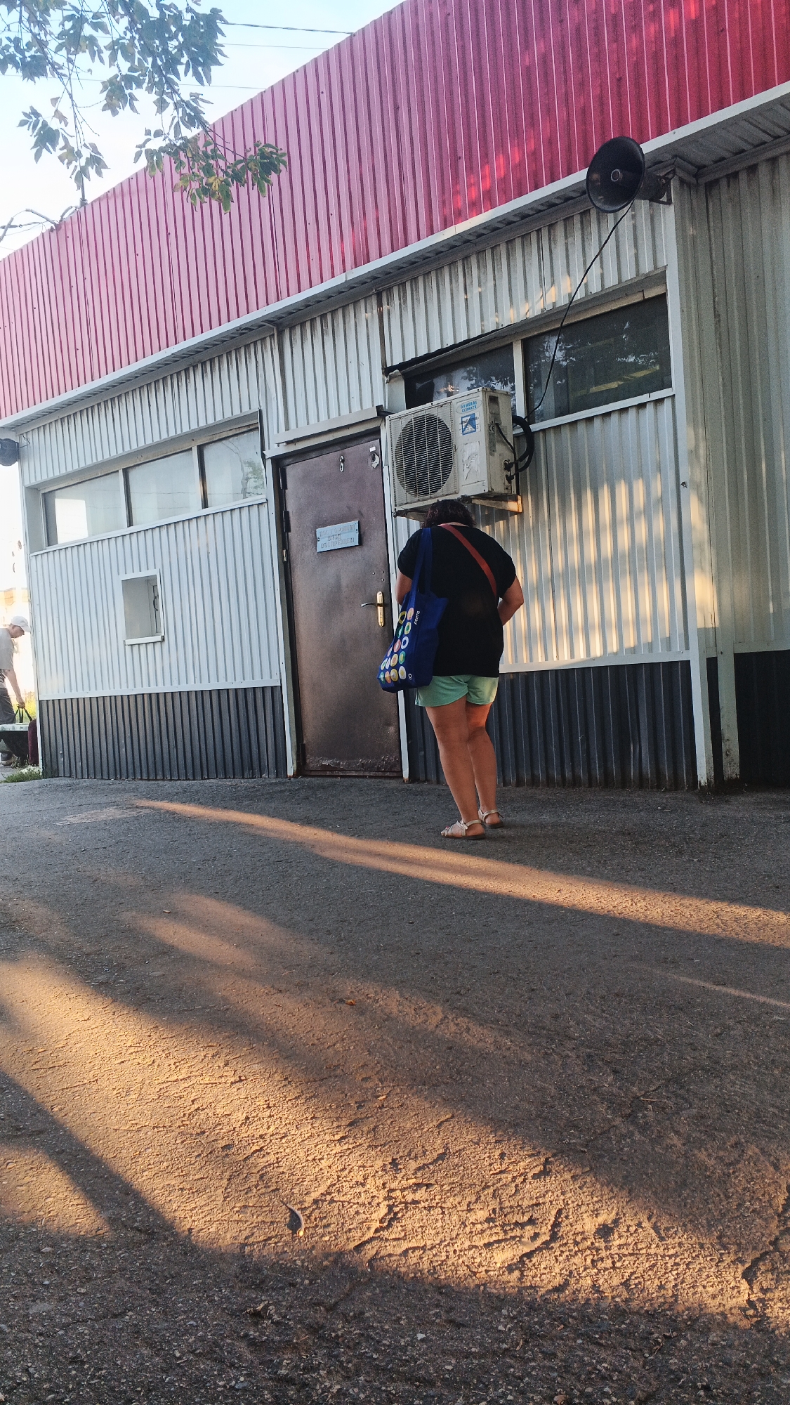 The image shows a woman standing in front of a corrugated metal building. The building has a red corrugated metal roof and a section of white corrugated metal siding. There is a door on the building with a sign above it. An air conditioning unit is mounted above the door. The woman is wearing a black t-shirt, light green shorts, and sandals. She is carrying a blue bag with colorful circular patterns over her shoulder. The ground in front of the building is asphalt with shadows cast across it, indicating the time of day is late afternoon or early evening, with sunlight coming from the left. The scene appears to be in a commercial or industrial area. This location is identified as being in Izhevsk, Russia.