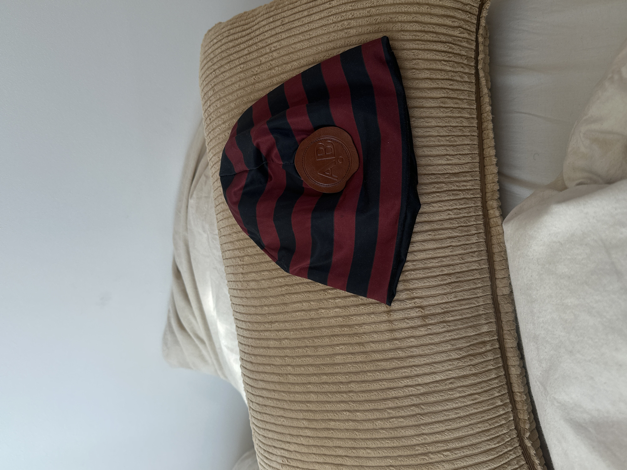 A red and black striped hat with a brown leather logo patch is lying on a beige pillow. The location is not discernible from the image.