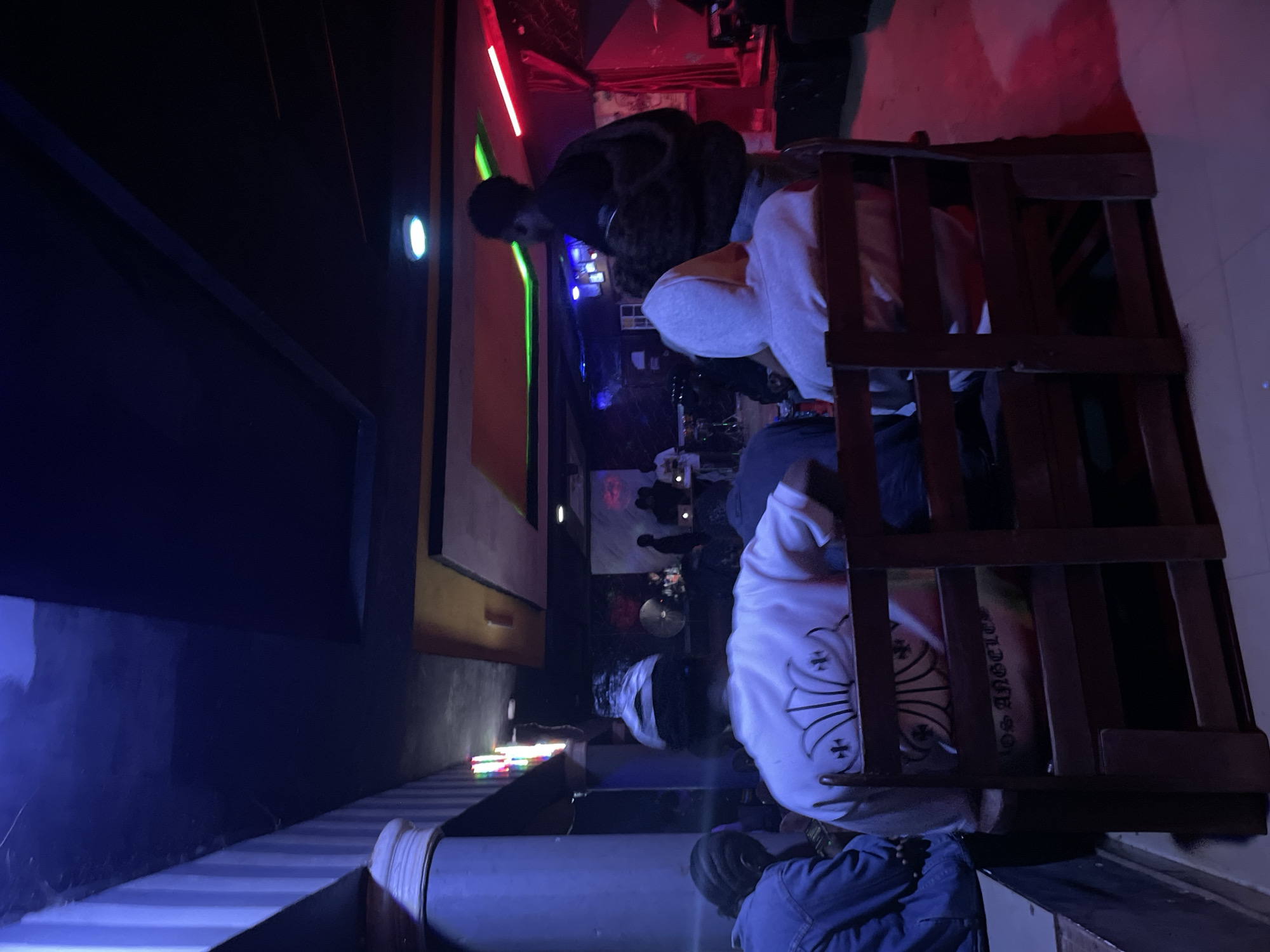 The image depicts an indoor scene, likely a club or performance venue, illuminated by an array of colored lights, including blue, green, and red, creating a vibrant and atmospheric setting. In the foreground, several individuals are seated on wooden chairs, facing away from the camera, observing an event happening in the background. One person in the center wears a white sweatshirt with a floral design and crosses. To the left, another person is visible from the back, wearing a denim jacket. The background features a stage area with musical instruments and several people standing. The walls are decorated with vertical striped patterns and abstract artwork, while the ceiling is adorned with recessed lighting and geometric paneling. The overall ambiance suggests a lively gathering, possibly a music performance or cultural event. This scene is located in Durumi, Nigeria.