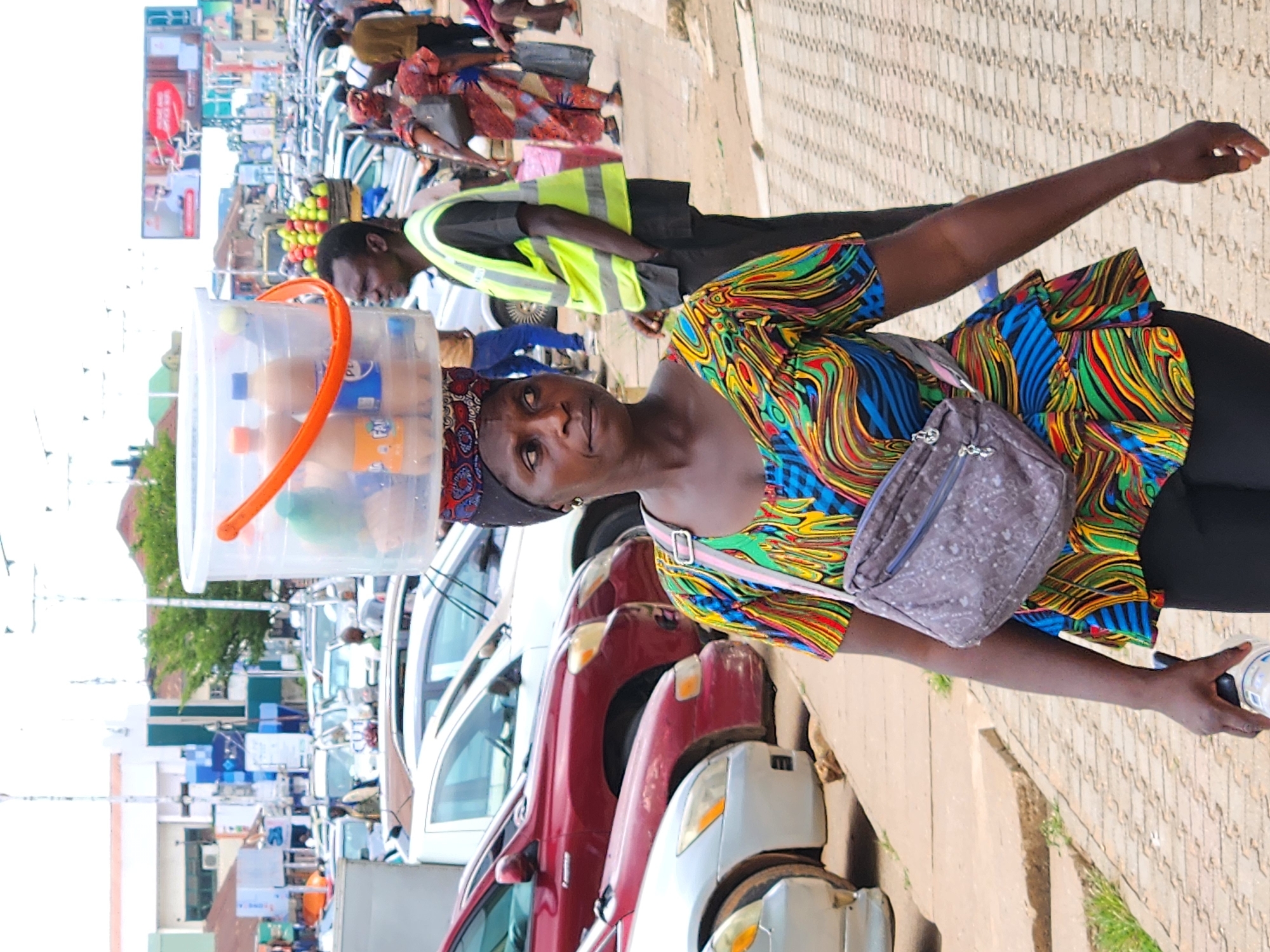 A woman is walking on a sidewalk in a city, carrying a large plastic bucket of drinks on her head. She is wearing a colorful, patterned shirt and black pants, with a grey fanny pack across her chest. She is holding a bottle in her right hand. Behind her, several cars are parked along the street, and other people are visible walking in the background. The environment appears to be a busy urban area with buildings and signage visible in the distance. The lighting suggests it is daytime.