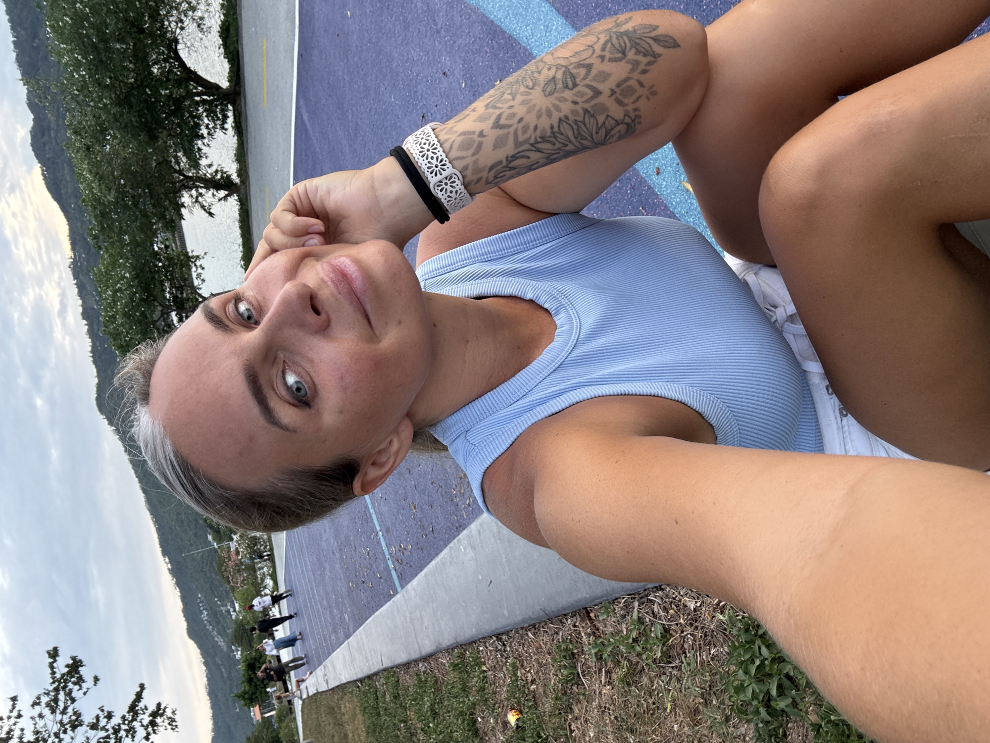 In a close-up, the subject, a woman with blonde hair and striking blue eyes, is captured from the chest up, positioned on a purple and blue paved surface, likely a recreational area. She is wearing a light blue ribbed tank top and white shorts. Her right arm rests on her knee, displaying a forearm tattoo of intricate floral patterns. A black hair tie and a white decorative bracelet adorn her wrist. Her left hand is gently touching her chin as she looks directly at the camera with a soft smile. In the background, a body of water is visible, bordered by lush green trees and a mountainous landscape. Several people are seen in the distance, some appearing to be walking or engaged in activities on a paved path. The sky is overcast with a hint of twilight. The location is identified as a street in Ko Samui District, Thailand.