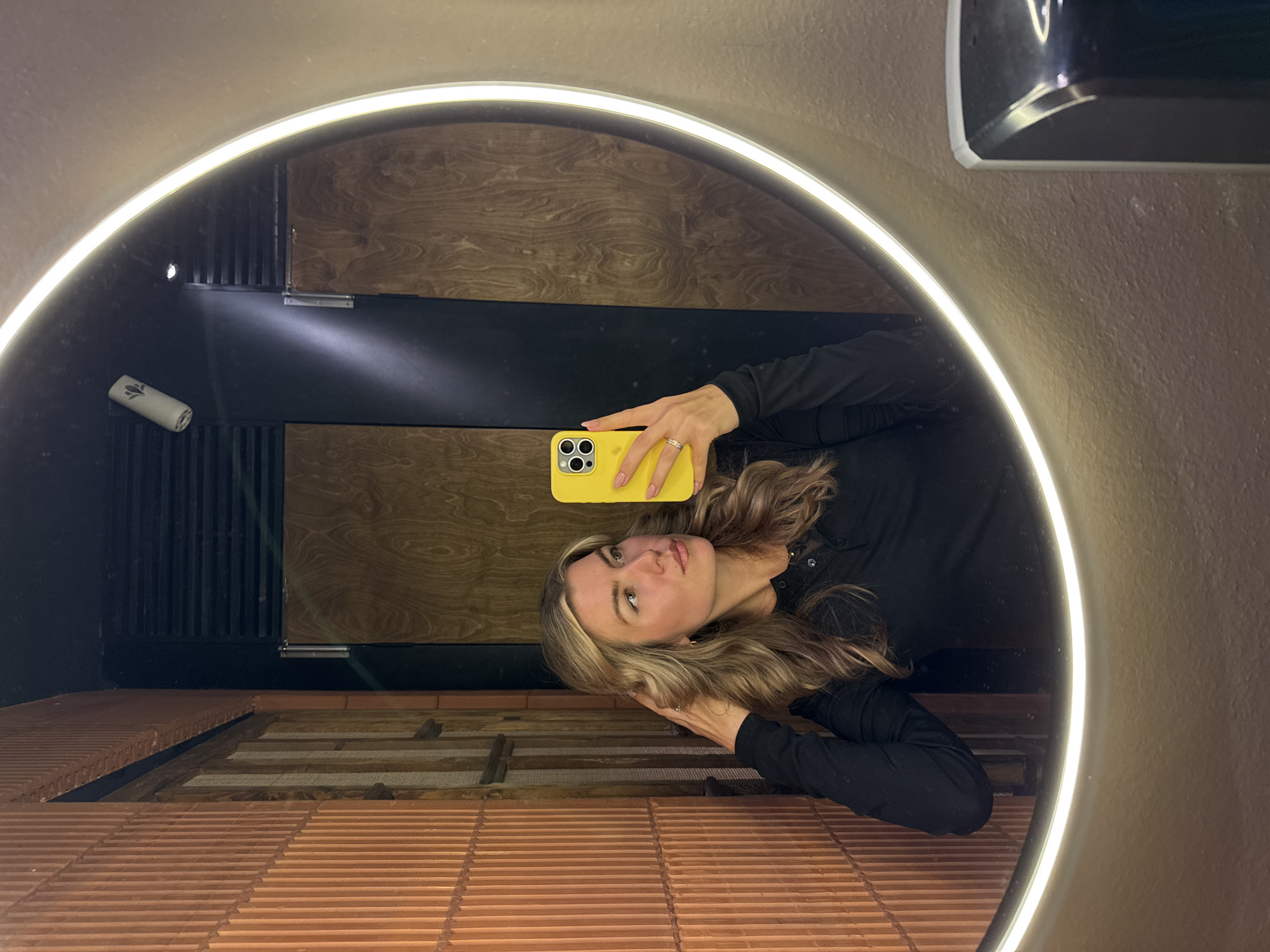 The image shows a woman taking a selfie in a round, illuminated mirror. She is holding a yellow iPhone in her right hand. She has blonde hair and is wearing a black long-sleeved shirt. The background reflected in the mirror shows a tiled floor, a black section with vertical lines, and a wooden wall. Above the mirror is a hand dryer mounted on the wall. The location appears to be a bathroom or restroom. The city and state are Unknown, Unknown.