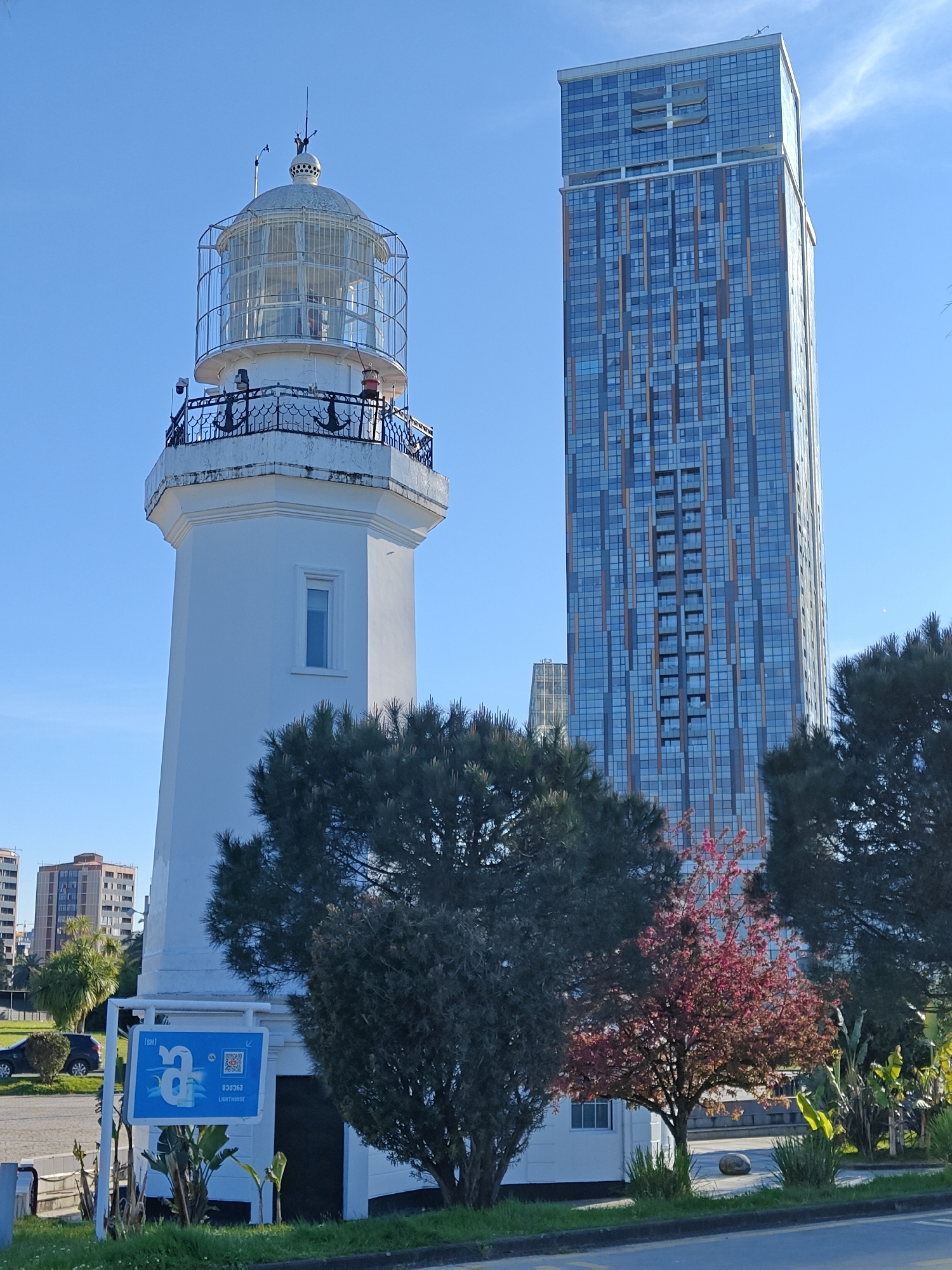 The image shows a lighthouse and a modern skyscraper in Batumi, Georgia. The lighthouse is white, cylindrical, and capped with a glass enclosure. A black iron railing with anchor motifs surrounds the viewing platform. The building is tall, with a blue glass facade patterned with horizontal and vertical bands. The sky is clear blue. Bushes and a small tree with pink flowers fill the space between the structures. A sign with the word "Lighthouse" in English and Georgian stands in front of the lighthouse. A road with a curb is in the foreground.