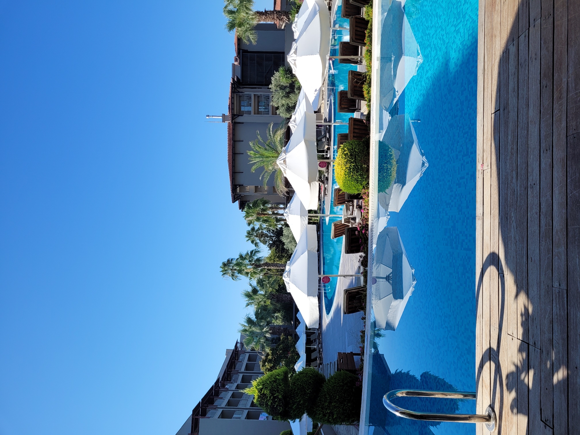 The image depicts a resort swimming pool area on a clear, sunny day in Manavgat, Turkey. A large, bright blue swimming pool with a tiled bottom occupies the center of the frame. Several white umbrella-shaped sunshades are positioned around the pool, with some casting reflections on the water's surface. Lounge chairs are visible behind the sunshades, suggesting a recreational area. In the background, a multi-story hotel building with balconies and a reddish-brown tiled roof is prominent, flanked by lush green trees and palm trees. The foreground features a wooden deck with a metal handrail partially submerged in the water, hinting at the entrance to the pool. The overall scene conveys a peaceful and inviting atmosphere of a holiday resort.