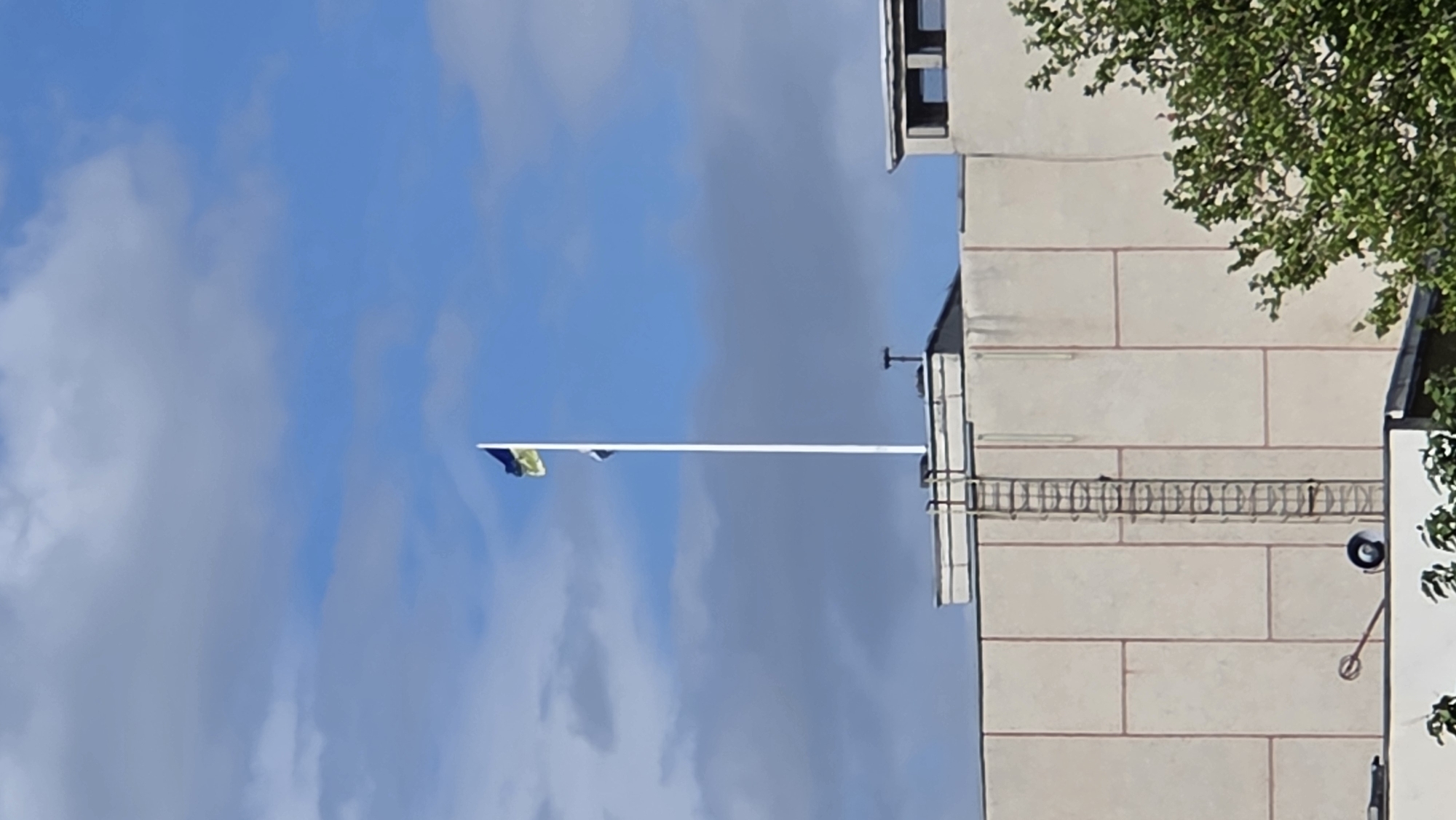 A Ukrainian flag, blue on top and yellow on the bottom, flies from a tall white flagpole mounted on the rooftop of a multi-story building. The building has a light-colored facade with vertical and horizontal paneling. A metal ladder is affixed to the side of the building, extending from the roof downwards. The sky is blue with scattered white clouds. Trees with green foliage are visible on the right side of the image. The location is the city of Pivdenne, Ukraine.