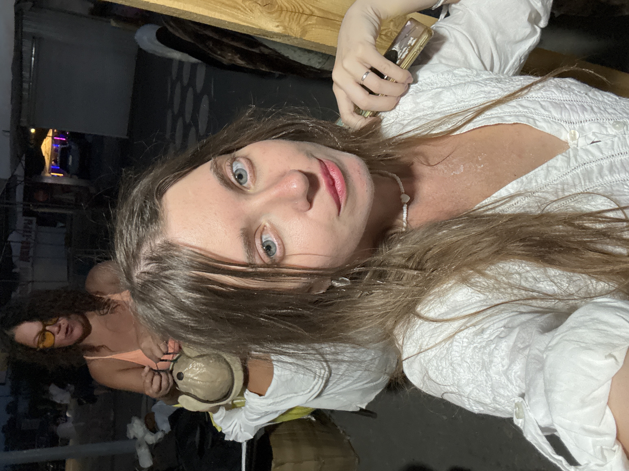 In Sol-Iletsk, Russia, a woman with long, light brown hair and light blue eyes, wearing a white blouse with embroidered details and a white heart-shaped beaded necklace, is in the foreground. She is holding a rectangular golden object, possibly a compact or a small electronic device, in her right hand. A thin silver ring is visible on her right ring finger. Her skin appears flushed, and there are visible marks on her chest.

Behind her, a child is seated, facing away from the camera. The child is wearing a white long-sleeved shirt and a light brown hat with ear flaps. To the left of the child, another woman with dark curly hair, wearing a coral-colored tank top and yellow-tinted glasses, is visible. She appears to be handling something with her hands.

The environment suggests an outdoor setting at dusk or night. In the background, there are structures resembling market stalls or outdoor seating areas, with some colorful lights visible, indicating a public or commercial space. The ground appears to be paved.