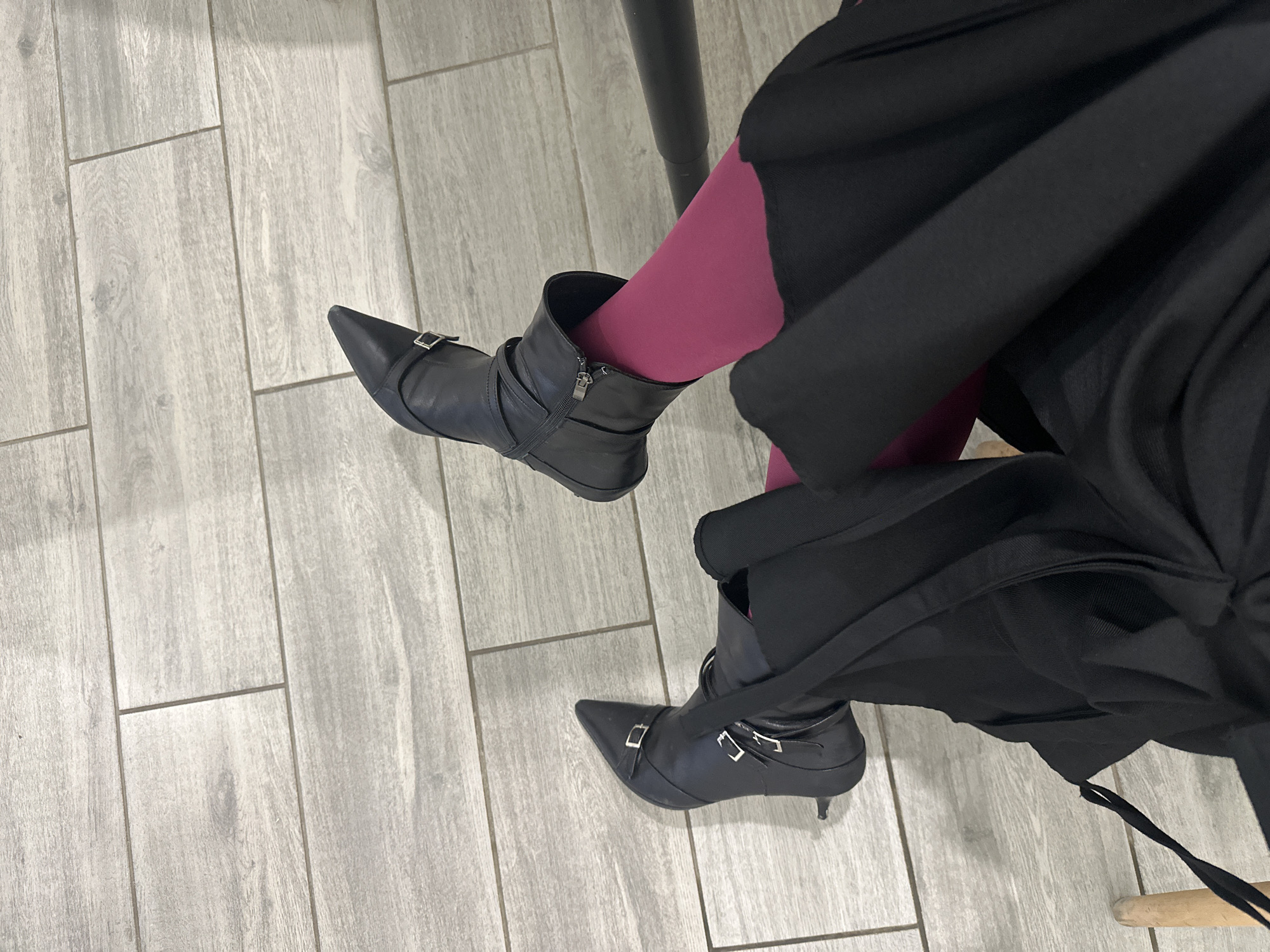 The image shows a person wearing pointed black heeled boots, magenta tights, and a black skirt. The boots have buckles and zippers. The person is sitting on a chair with a dark leg, and the background is a light grey wood-patterned floor. The scene is indoors. There is no indication of a specific location in Moscow, Russia.