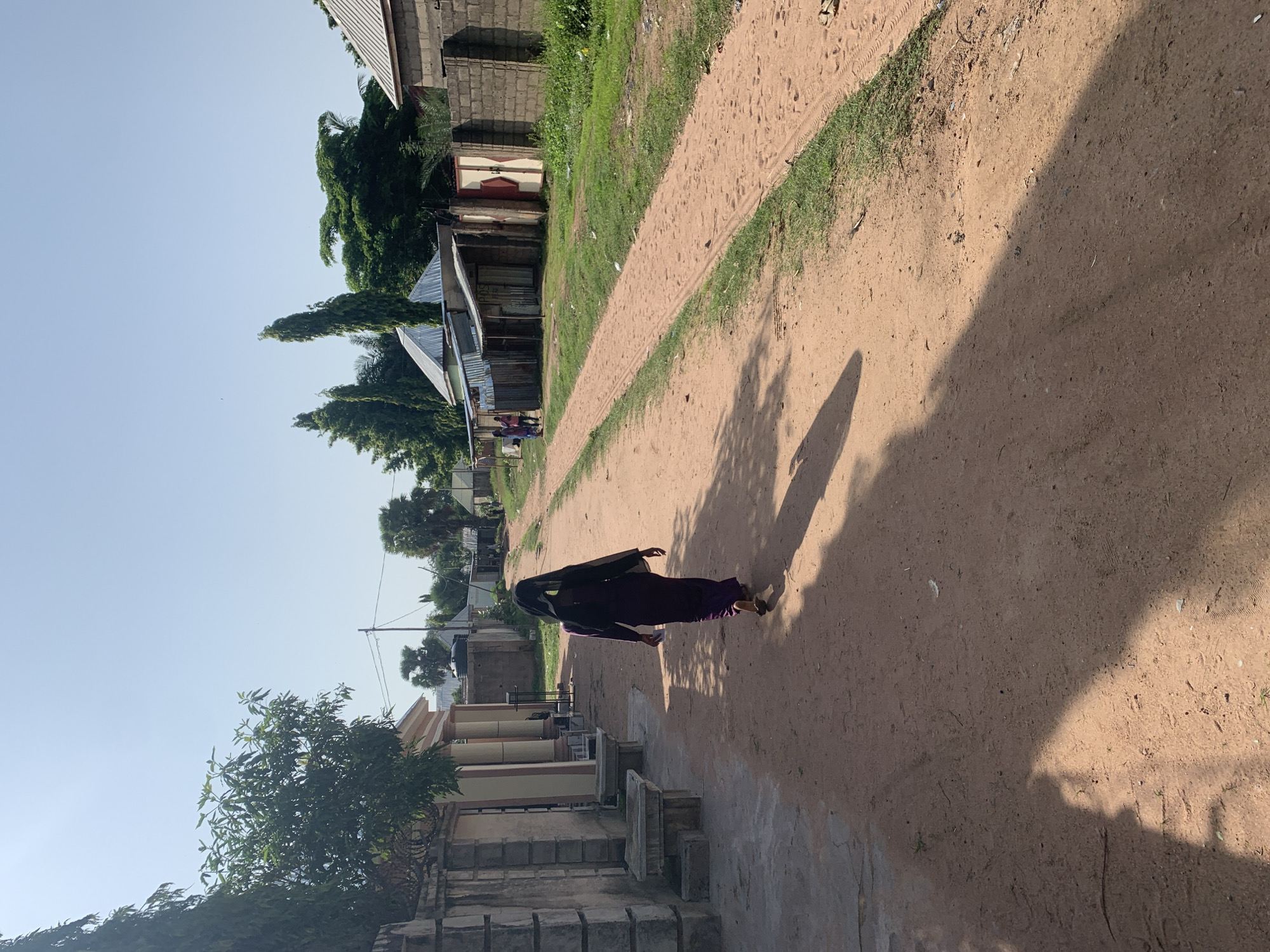 A dirt road passes through a residential area in Jalingo, Nigeria, under a clear sky. On the left side of the road, a person wearing a dark purple dress and a black headscarf walks away from the camera. They are casting a long shadow on the dusty ground. Adjacent to the road on the left is a wall with large pillars and what appear to be benches. On the right side of the road are various buildings, including some with corrugated metal roofs and brick walls. Green grass grows along the edges of the road and in patches between the buildings. In the distance, more buildings and trees are visible, and a few other people can be seen further down the road. The overall scene depicts a sunny day in a seemingly quiet neighborhood.