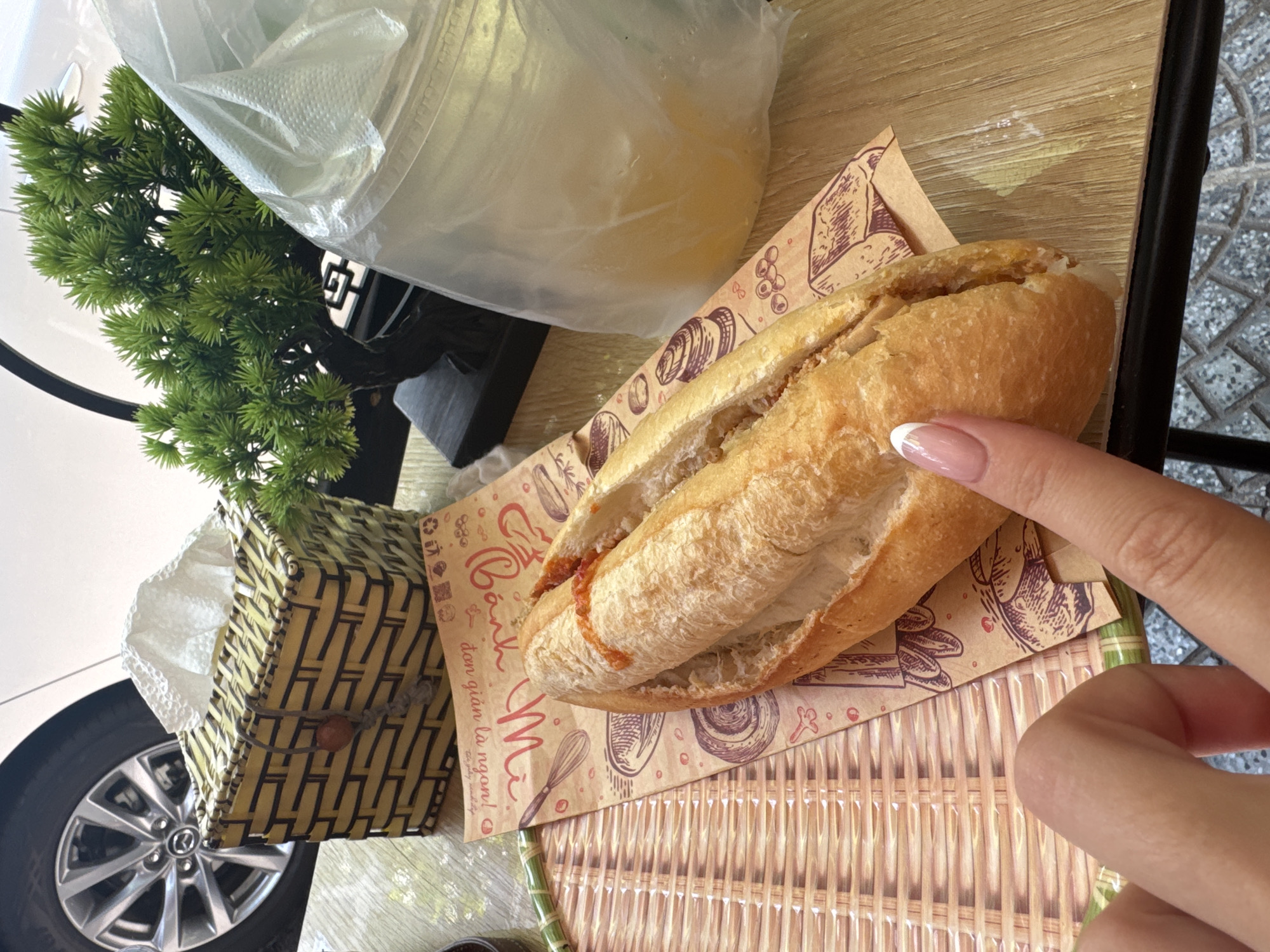 A close-up, slightly high-angle shot shows a partially eaten bánh mì sandwich on a printed paper wrapper. A hand with manicured nails points toward the sandwich. The sandwich is on a woven tray. Beside the tray is a tissue box in a woven container. The background also includes a small plastic plant, a clear plastic bag containing contents that are out of focus, and what appears to be a car tire. Based on the packaging on the sandwich, it is likely in Đà Nẵng, Vietnam. The scene appears to be in a vehicle.