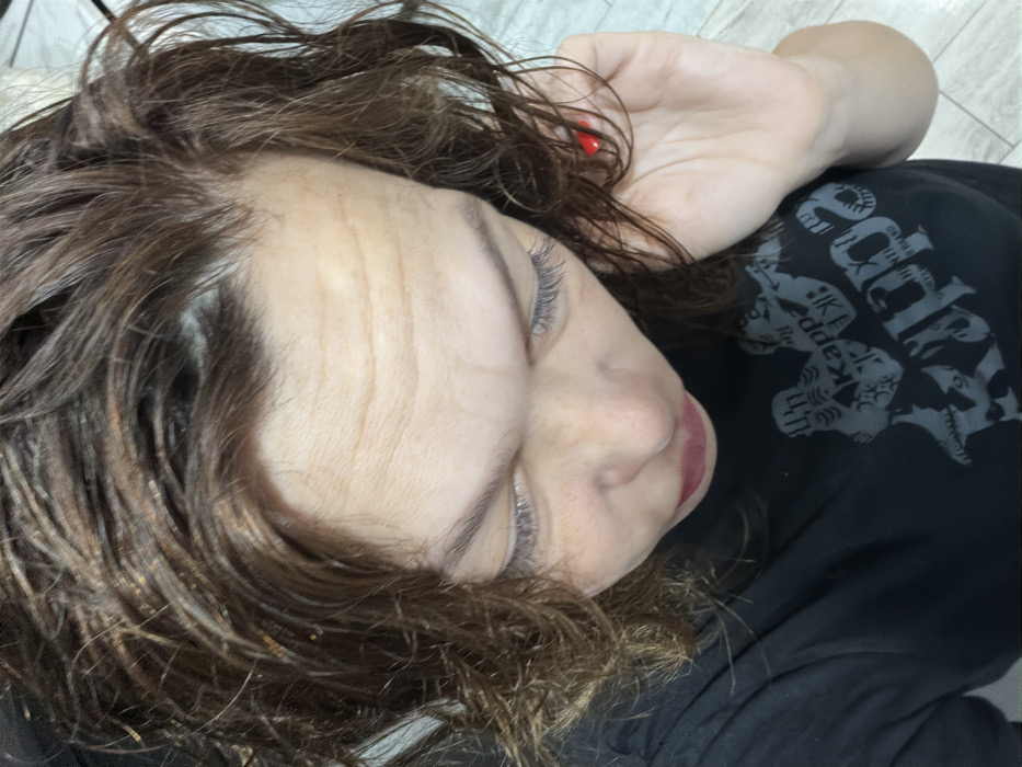 The scene is located in the city of Chaikovsky, Russia. The image displays an adult female observed from an overhead perspective, looking downwards. She has wavy, medium-brown hair that appears wet or damp, featuring lighter streaks. Her forehead exhibits multiple horizontal wrinkles. Her eyes are downcast and partially visible, with noticeable eyelashes. Her lips are closed and appear colored with a reddish-pink product. She is wearing a dark, short-sleeved t-shirt adorned with a grey graphic print, identifiable as the Kappa brand logo accompanied by stylized figures and text. Her right hand, featuring a red-painted thumbnail, is positioned near the right side of her head, gently touching her hair. The background is out of focus, showing a light-colored, uniform surface, possibly an interior floor made of tile or laminate.