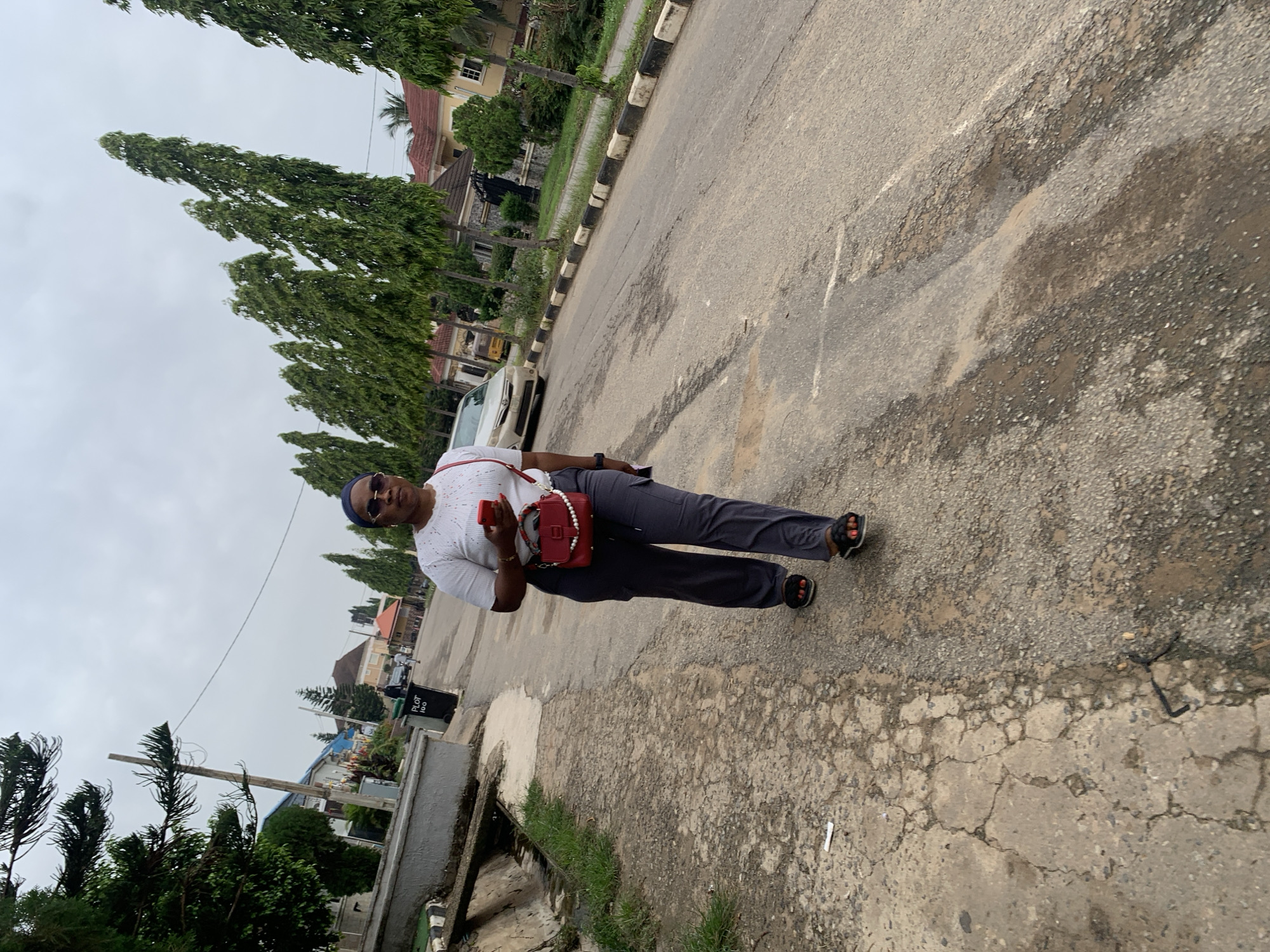 A full-body, slightly low-angle outdoor shot captures a Black woman walking on a paved street in a residential area of Abuja, Nigeria. She is positioned in the lower-center of the frame, facing the camera and slightly to her left. She wears a white t-shirt with a textured pattern, dark grey cargo pants, black sandals, a dark blue headscarf, and sunglasses. A red crossbody purse with pearl accents is slung over her right shoulder. Her left hand holds a red smartphone.
The street is uneven with visible cracks and patches. To the left of the street, a drainage channel runs alongside a low concrete wall topped with green grass and a thin tree. Behind the woman, a row of tall, slender evergreen trees lines the street. Several residential buildings with red-tiled roofs are visible in the background, along with parked cars and a yellow autorickshaw. The sky is overcast and cloudy. The scene suggests an ordinary day in a suburban neighborhood of Abuja.
