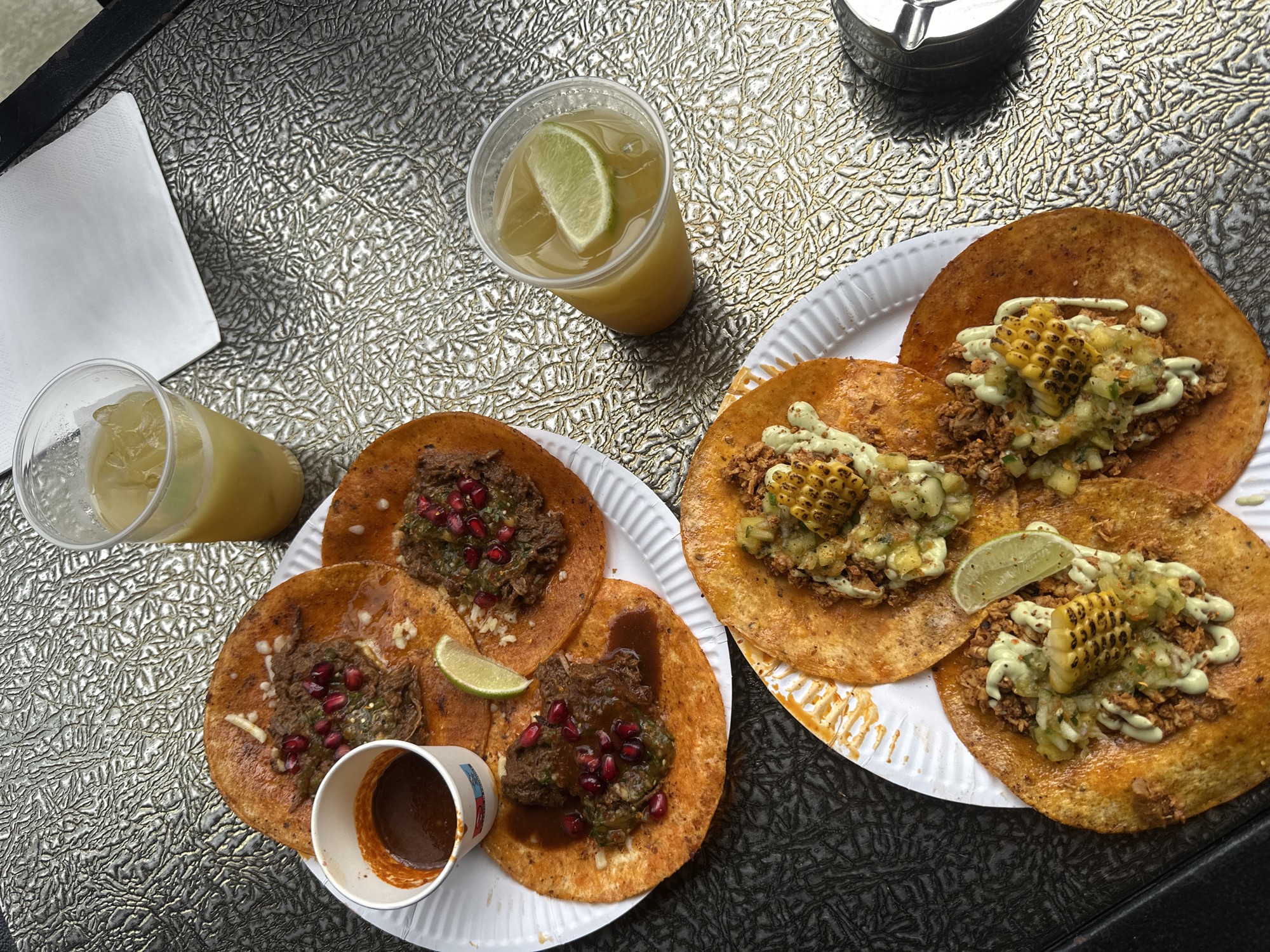 The image shows two paper plates of tacos and two drinks on a reflective surface. The tacos on the left plate are topped with meat, pomegranate seeds, and cilantro, and there is a small cup of sauce. The tacos on the right plate have meat, grilled corn, and a green sauce. The drinks are in plastic cups with lime wedges. A white napkin is visible on the left, and a black object is in the upper right corner. There is no indication of the location being in London, United Kingdom, or any people present in the scene.