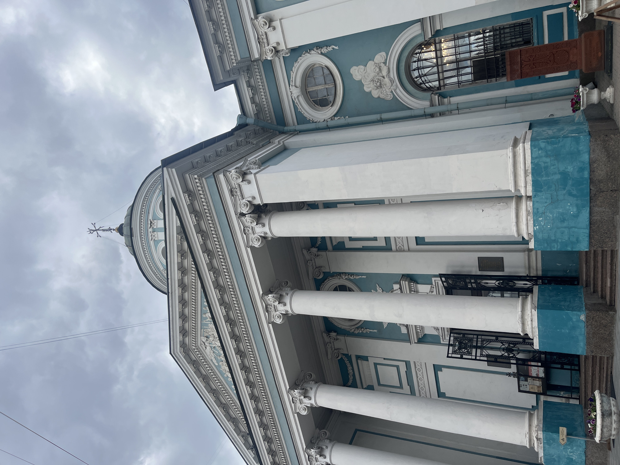This image depicts the exterior of a building with classical architectural elements, likely a church or a public building, in Saint Petersburg, Russia. The building features a prominent facade with large white columns topped with Corinthian capitals. The walls are painted in a striking combination of white and turquoise blue. Above the columns, there is a decorative frieze with bas-relief sculptures. A dome with a cross on top is visible in the background, characteristic of Orthodox Christian churches. The sky is overcast, with dramatic clouds suggesting it might be a cloudy day. In front of the entrance, there are black metal gates and steps. Planters with flowers are placed on either side of the entrance. To the right of the entrance, a tall, reddish-brown stone monument, possibly a khachkar (a traditional Armenian stone cross), stands near a large arched window with a metal grate. The overall impression is one of grandeur and historical significance, set against a subdued sky.