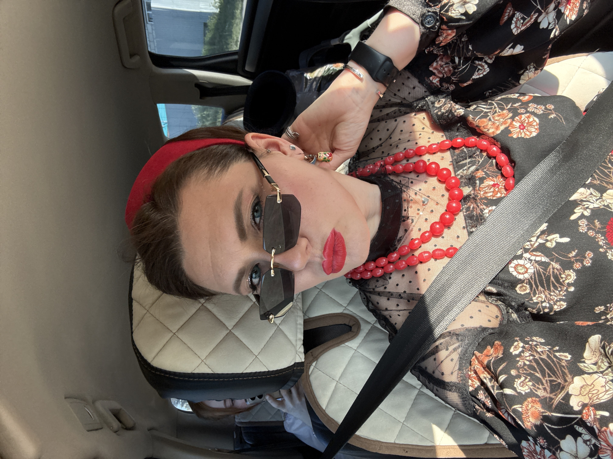 A woman with red lipstick, sunglasses, and a red headband is in a car. She is wearing a black floral print shirt and a red beaded necklace. She has a black watch on her wrist. She is wearing a seatbelt. The headrest of the car seat is tan with a black border. The location is likely Chaikovsky, Russia.
