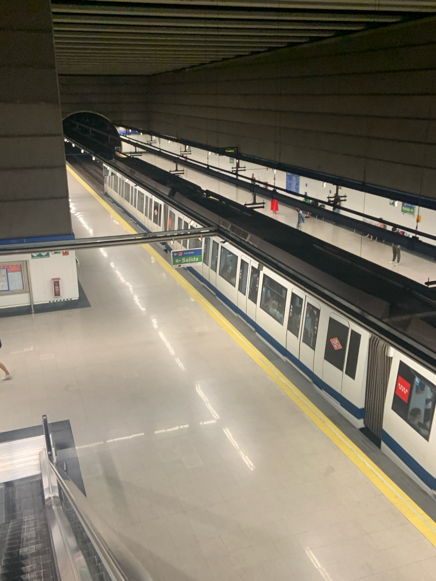 The image depicts a view from above of a modern underground metro station. A white train with blue trim is stopped at the platform, which is marked with a yellow safety line. Several doors on the train are visible, some with tinted windows showing passengers inside. Across the tracks, another platform is visible, also with a yellow safety line. On this platform, there are several people standing and sitting, some waiting for a train. Signs are posted, including a green and white sign with an arrow pointing left and the word "Salida" (Exit), indicating an exit route. A small white building with a red fire extinguisher mounted on the exterior is also present on this platform. The station's ceiling is dark and ribbed, with overhead lighting. In the background, a tunnel entrance for the train tracks is visible. The floors of the station appear to be tiled and reflective, with some white lines painted on them. The scene is likely within the Alcorcón Metro system in Alcorcón, Spain, given the signage and train design which is characteristic of the Madrid Metro.