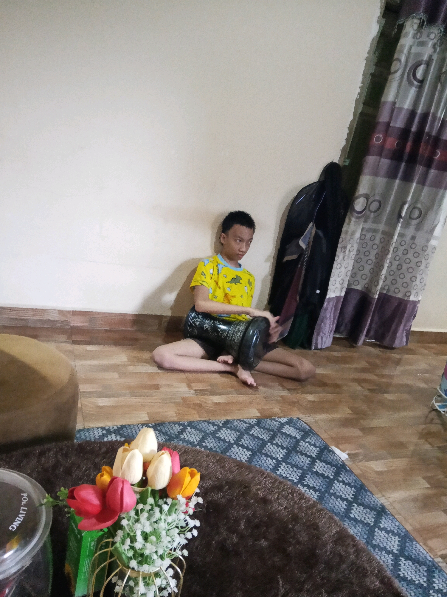 The image depicts an indoor scene, possibly a living room, in Lubuklinggau, Indonesia. A young boy, wearing a yellow t-shirt with blue accents and dark shorts, is seated cross-legged on a wooden floor. He is holding a traditional drum, possibly a darbuka, resting it on his lap. His left hand is on top of the drum, and his right hand is positioned as if ready to play.
The background features a light-colored wall, with a wooden skirting board running along its base. To the right, a curtain with a pattern of concentric circles in shades of purple, grey, and white hangs from a rod. In front of the curtain, a dark garment, possibly a jacket or coat, is draped over something.
In the foreground, a brown, textured rug is partially visible, partially covered by a patterned blue and white rug. To the left of these rugs, a round, beige ottoman is present. Further left, a part of a glass container with a metal lid is visible, and text that appears to read "POL LIVING" is visible on the lid. In front of the rugs, a bouquet of artificial tulips in red, yellow, and cream colors is arranged in a gold wire vase, with small white artificial flowers interspersed. The lighting suggests an indoor environment, likely during the day.