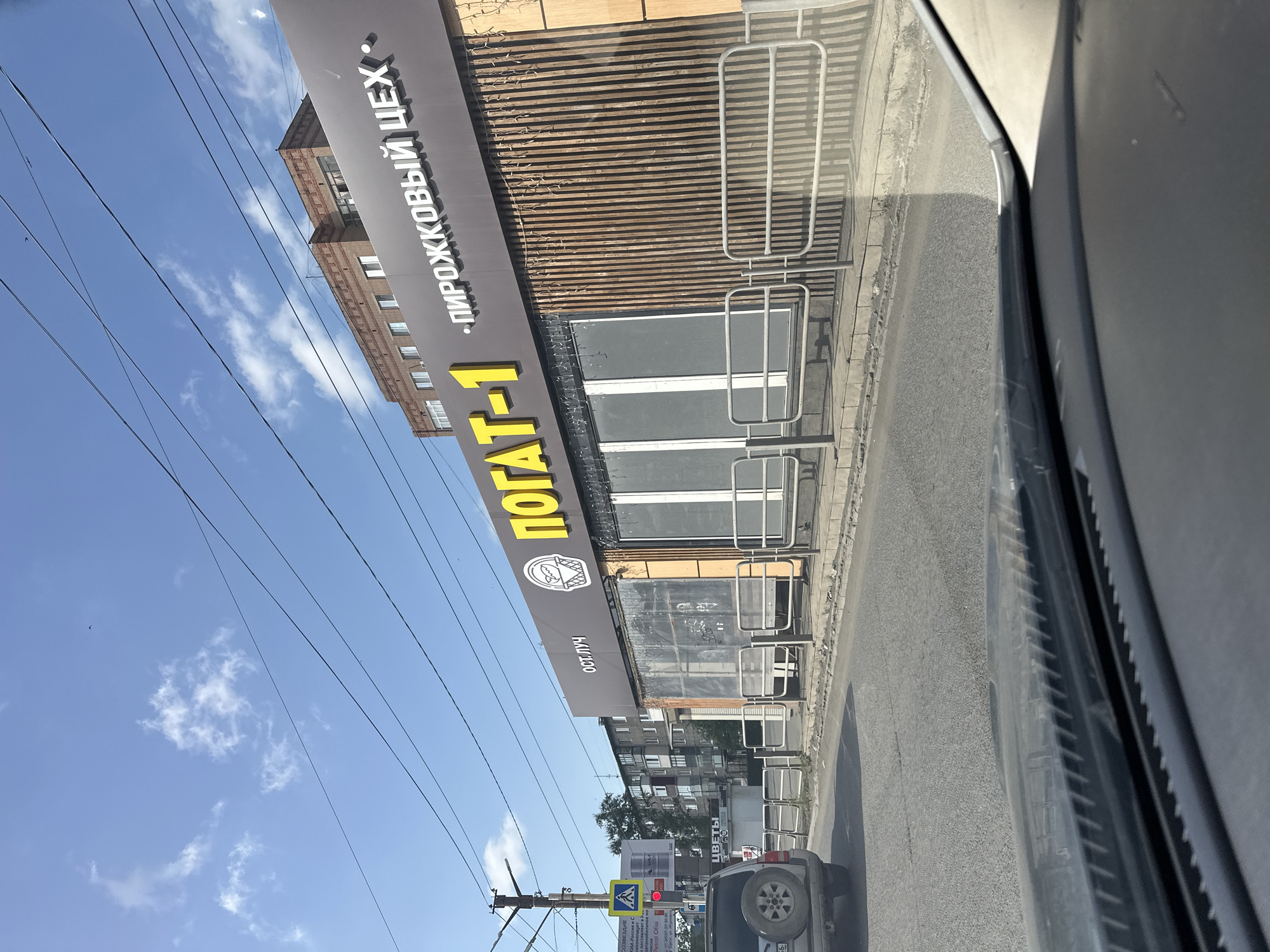 This is a photograph taken from inside a vehicle looking forward at a street scene in Orsk, Russia. The building on the right side of the image features a large sign with yellow lettering that reads "ПОГАТ-1" and "• ПИРОЖКОВЫЙ ЦЕХ •", indicating a pie shop or bakery. To the left of this sign, there is a smaller sign that says "ОСТ. ЛУЧ". The building itself has a modern facade with vertical wooden slats and large, covered windows. A metal fence runs along the sidewalk in front of the building. In the foreground, the dashboard of the vehicle is visible. Further down the street, there are apartment buildings, trees, and utility poles with traffic signs. A silver SUV is parked on the left side of the road, and the general environment appears to be a sunny day with a clear blue sky and some scattered clouds. The presence of the sign in Russian clearly places the location in Russia.