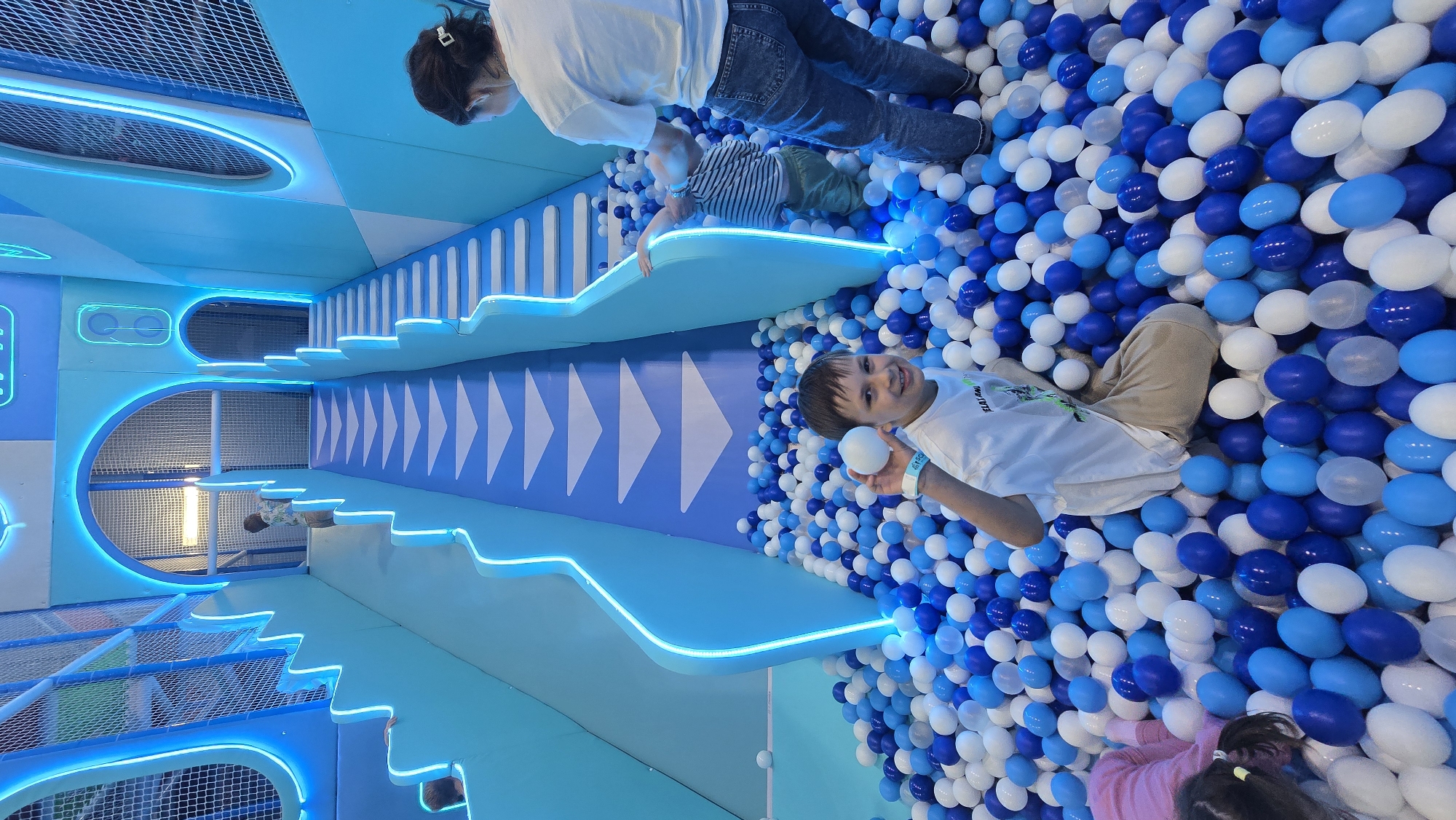 This image captures an indoor children's play area, specifically a multi-level ball pit and slide structure, located at Belani Land in Izhevsk, Russia. The environment is characterized by a bright, modern design predominantly featuring shades of blue (light blue, medium blue, dark blue) and white, with prominent LED strip lighting illuminating the edges of the structures in a cool white-blue hue. In the foreground, a boy approximately 8-12 years old, wearing a white short-sleeved t-shirt and light brown shorts, is seated in the ball pit. He is smiling and looking directly at the camera, holding a single white plastic ball in his right hand. A light blue wristband, visibly bearing text including "BELANI.LAND IZHEVSK," is on his left wrist. The ball pit itself is densely filled with plastic balls in white, light blue, and dark blue colors. To the immediate left of the foreground boy, a younger child, estimated to be 3-6 years old, is partially submerged in the ball pit, lying face down. Only the back of their head with dark hair and a yellow hair tie, and parts of a pink long-sleeved top, are visible. In the mid-ground, to the right of the foreground boy, an adult, likely female, is standing in the ball pit. She is wearing a white t-shirt and blue denim jeans, with her dark hair pulled back. She is facing away from the camera, leaning over and attending to a smaller child, approximately 1-3 years old, who is wearing a striped top and green pants and is partially submerged in the balls next to a light blue structural element. The background features a large, multi-tiered play structure. On the left side, a wide slide or ramp descends into the ball pit, marked with a series of white downward-pointing arrow shapes on a darker blue surface, bordered by light blue panels with internal LED lighting. To its right, a corresponding staircase, also in light blue with LED-illuminated edges, ascends. Near the top of the left ramp, another child, estimated to be 3-6 years old and wearing a light-colored patterned shirt, is visible near an arched opening. Several arched passageways, lined with blue neon lighting, are present in the upper sections of the structure, some leading to areas enclosed by netting. The walls of the play area are a mix of light blue and white, with distinct horizontal lines and occasional oval-shaped design elements on the
