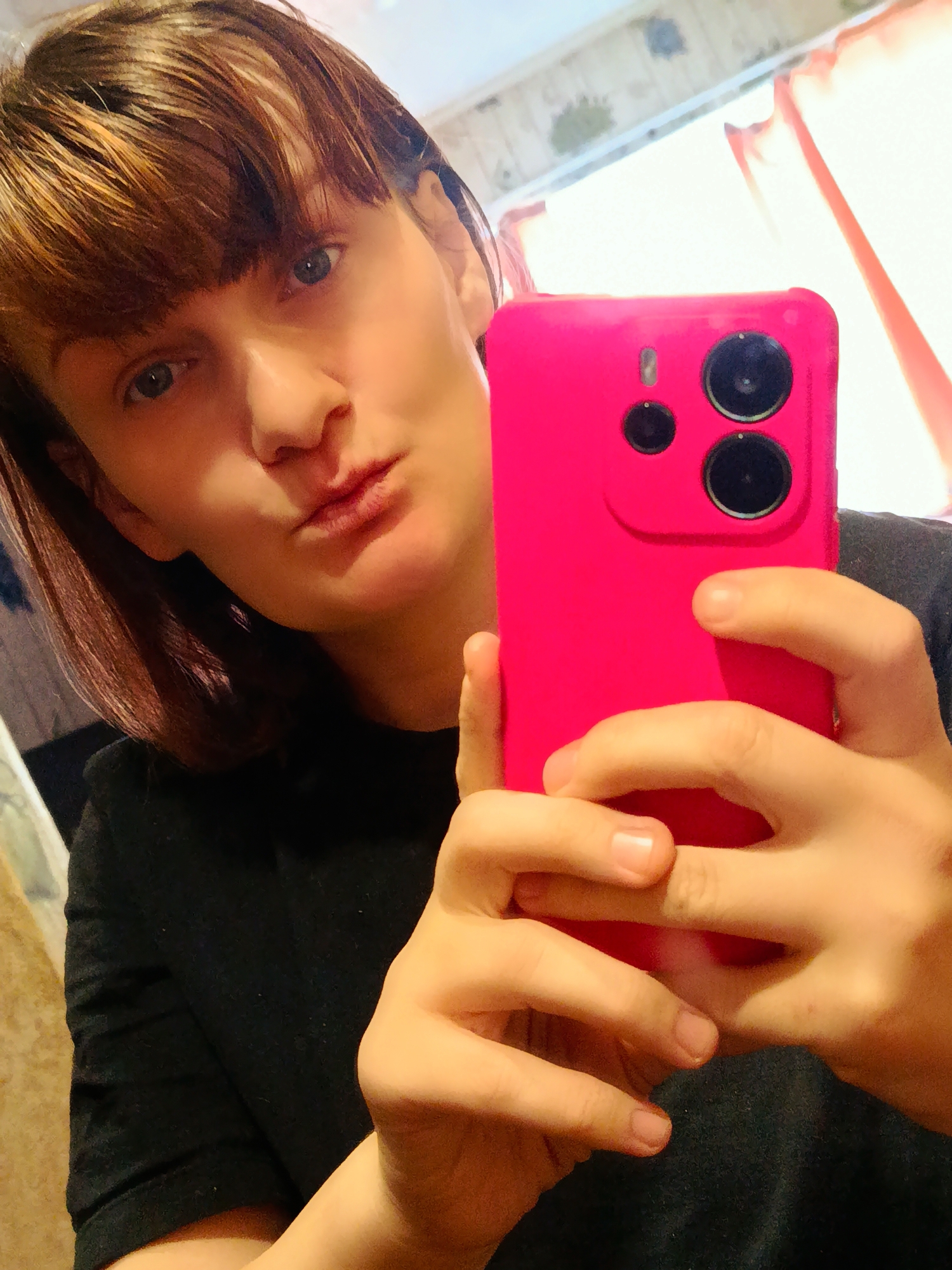 A woman with brown hair and blue eyes is taking a selfie with a hot pink cellphone. She is wearing a black shirt and puckering her lips for the camera. The background shows a room with light-colored walls and a window with red curtains. It is not possible to determine if the location is Фоки, Russia from the image.