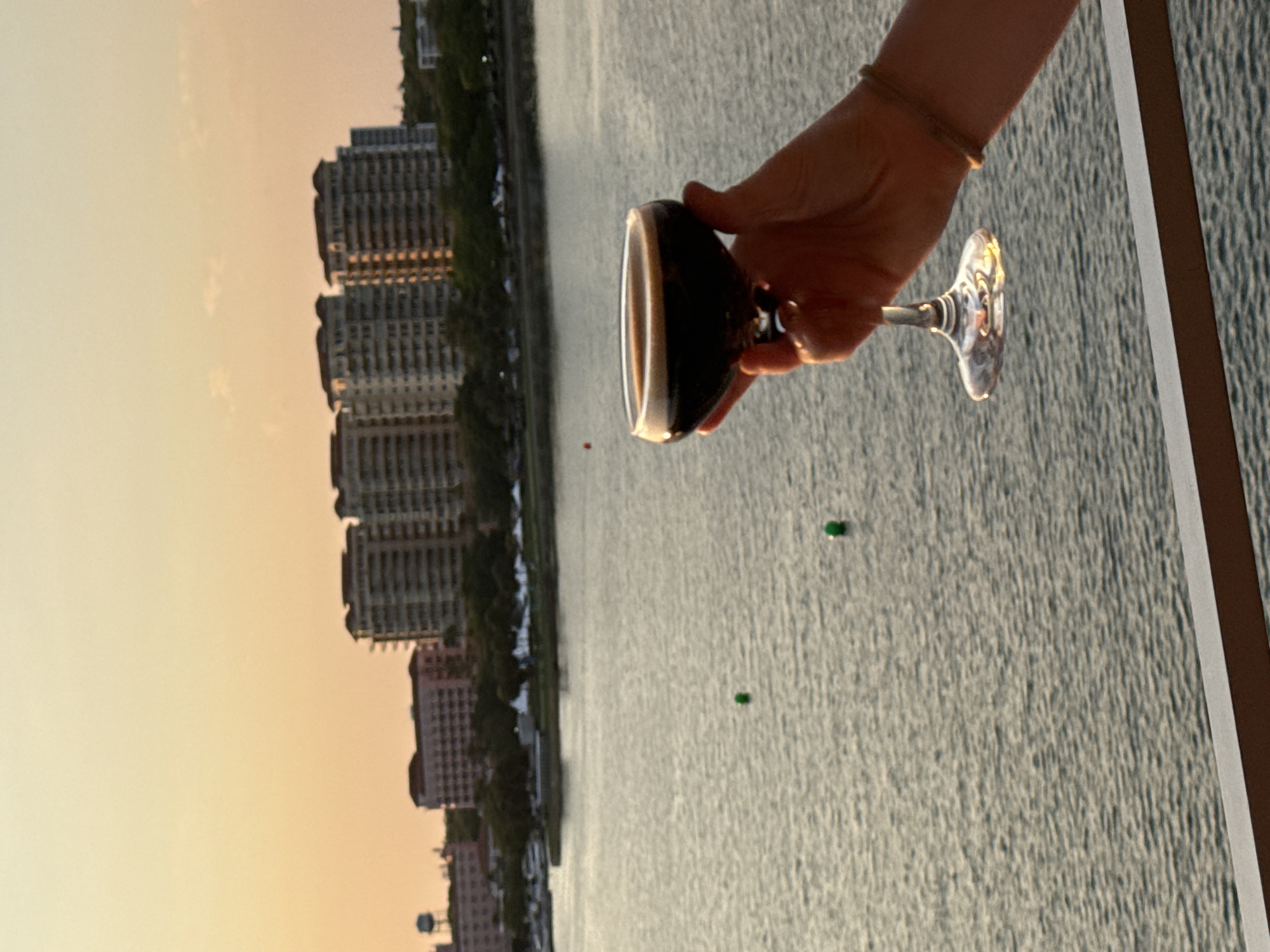 The image displays an outdoor scene at sunset in Saint Petersburg, Florida, United States, looking east-southeast over Tampa Bay towards the downtown waterfront.
In the lower right foreground, a person's left hand, adorned with a small ring on the ring finger and a light-colored bracelet on the wrist, holds a clear stemmed coupe glass. The glass contains a dark brown liquid topped with a thin layer of lighter-colored foam. The hand and glass are positioned above a dark brown railing that runs horizontally across the bottom of the frame.
The midground features the calm, rippled surface of Tampa Bay, reflecting the warm, golden light of the setting sun. Two green spherical buoys are visible floating on the water: one in the center-left and another further right and lower. On the opposite shore, a tree line and a marina with several docked boats are visible, along with multiple white canopy tents set up near the water's edge.
The background cityscape comprises various multi-story buildings under a clear sky transitioning from pale yellow to light orange. Several prominent modern residential or commercial towers are visible. To their left, the distinctive light pink Vinoy Renaissance St. Petersburg Resort building is identifiable. Further left, a dark-colored water tower stands against the horizon. The low-angle sunlight illuminates the western facades of the buildings.
