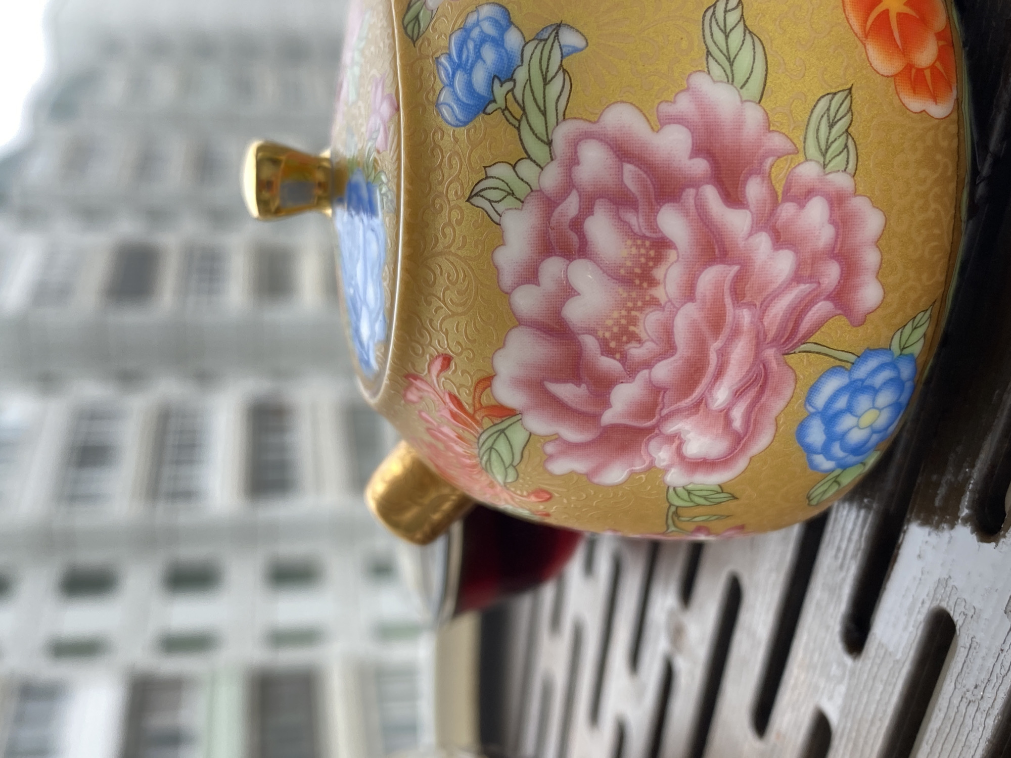 The image captures a close-up view of a decorative, likely antique, Asian-style teapot or covered container with floral patterns in pink, blue, and orange against a gold background. The teapot is situated on a dark, possibly wooden, latticed surface. In the blurred background, the outline of a building with multiple windows suggests an urban setting, possibly in Chaikovsky, Russia.