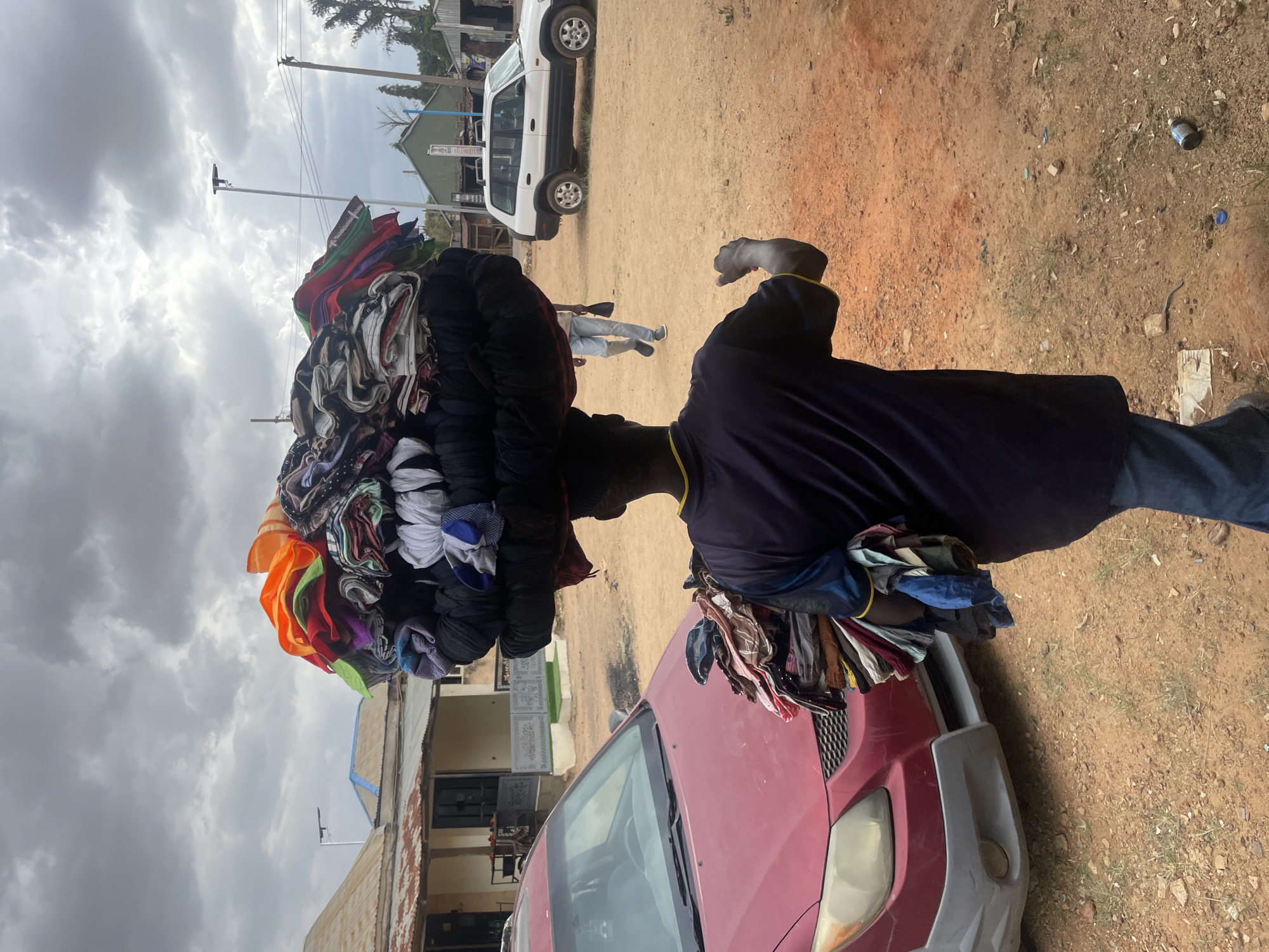 An outdoor scene depicts a male street vendor carrying a substantial stack of clothes on his head, walking on an unpaved, reddish-brown dirt road under an overcast sky. He is wearing a dark blue shirt with yellow trim and blue pants, with additional garments draped over his left arm.
In the foreground, a red car is partially visible to the vendor's left. Further back, another person in light-colored pants walks in the distance. A white SUV or jeep is parked in the mid-ground, and behind it, various buildings with corrugated metal roofs line the road, suggesting a semi-urban or rural setting. Utility poles and power lines are visible against the cloudy sky. The ground shows signs of scattered debris, including what appears to be a can and plastic items. The activity portrays daily commerce, typical of street vending in many regions. The time of day is daytime, indicated by the overall brightness despite the heavy cloud cover. No discernible text is visible in the image.