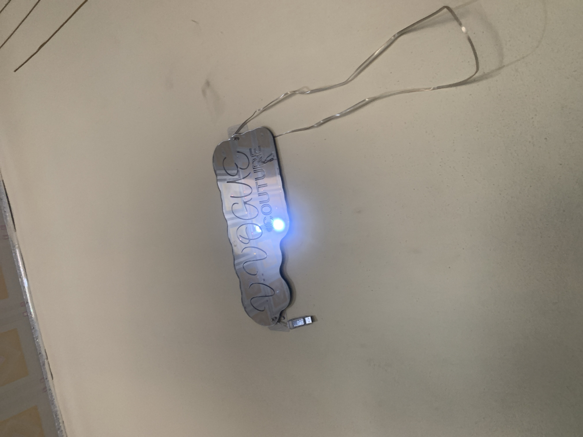 The image displays a reflective, cloud-shaped tag with the etched text "VOGUE COUTURE". A blue LED light illuminates the tag from behind, casting a glow. A small, silver USB drive is attached to the left side of the tag, with thin wires extending from the right side. The tag and wires are resting on a pale, textured surface, possibly a countertop or table, with some light and shadows visible. The background is blurred and appears to be an indoor environment, possibly a domestic or commercial setting. There are no people or animals visible in the image, and no specific location in Jauro Yinu, Nigeria can be identified.