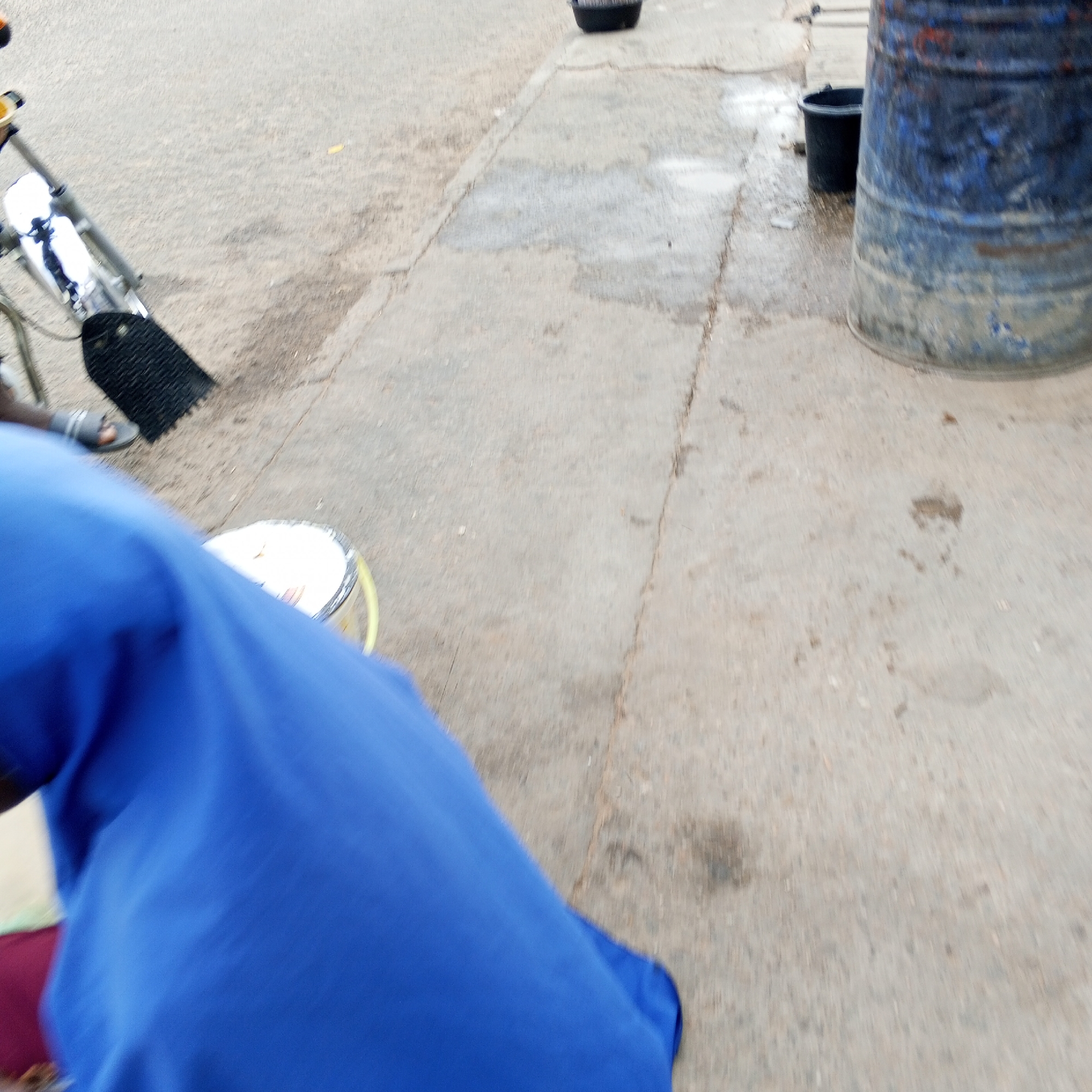 The image depicts a street scene in Arawa, Nigeria. In the foreground, a person's blue garment is visible, with a foot wearing a sandal resting on a black broom head. A yellow bucket containing a white substance is positioned near the person. In the background, a large, blue, metal barrel stands beside a smaller black bucket. The ground is paved with concrete slabs. The scene appears to be outdoors, likely on a street or walkway.